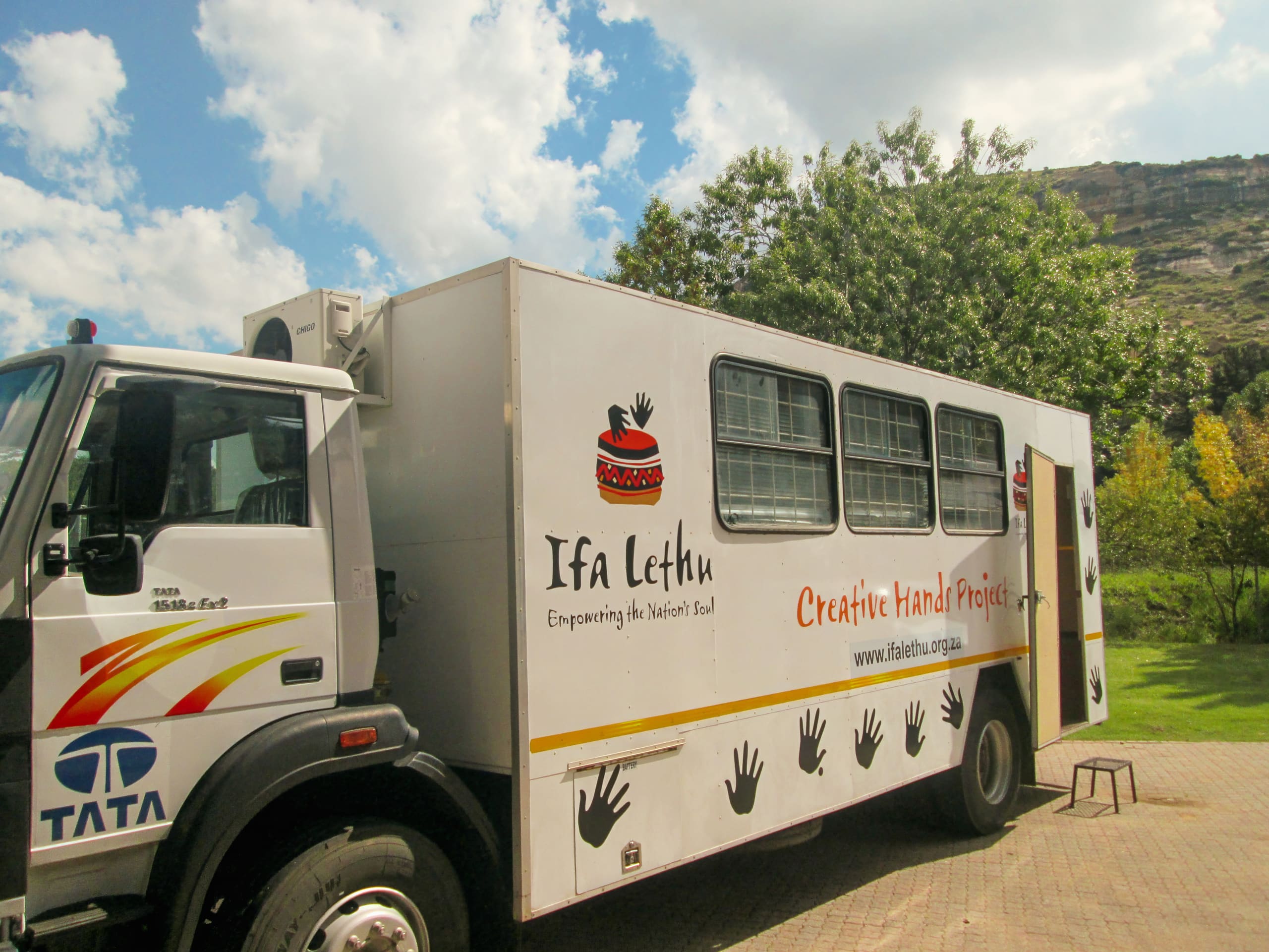 A large white lorry labelled “Ifa Lethu Creative Hand Project” is parked outdoors. The side of the lorry displays black handprints, text, and a logo with a raised hand and red bands. The sky is partly cloudy and trees and hills are visible in the background.