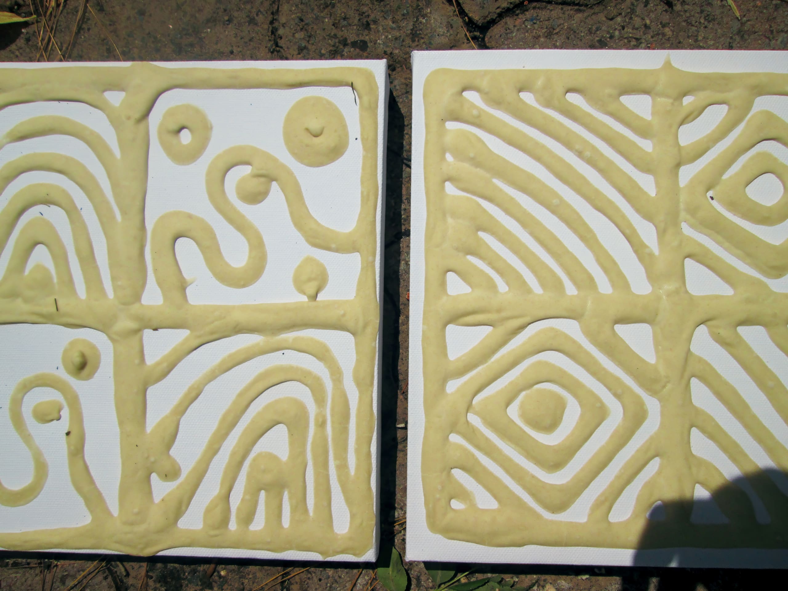 Two square canvases with raised, abstract patterns made of thick, beige paste. The left canvas features swirls, dots, and curved lines; the right canvas has diagonal lines forming diamond and oval shapes. Both rest on a rough stone surface.