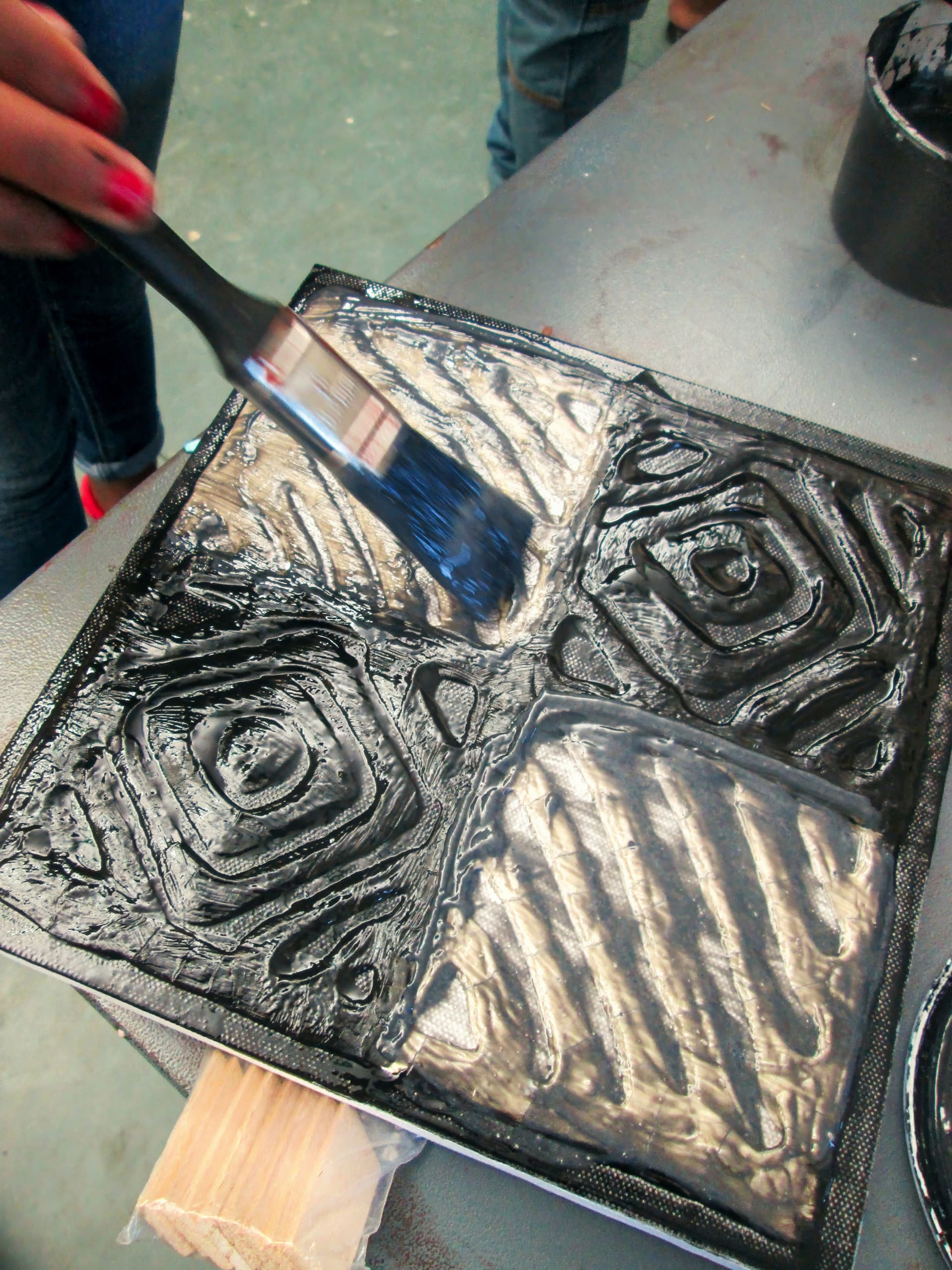 A hand with red nail varnish uses a paintbrush to apply black paint onto a textured, square tile with geometric patterns. The tile is on a work surface, and another person stands in the background.