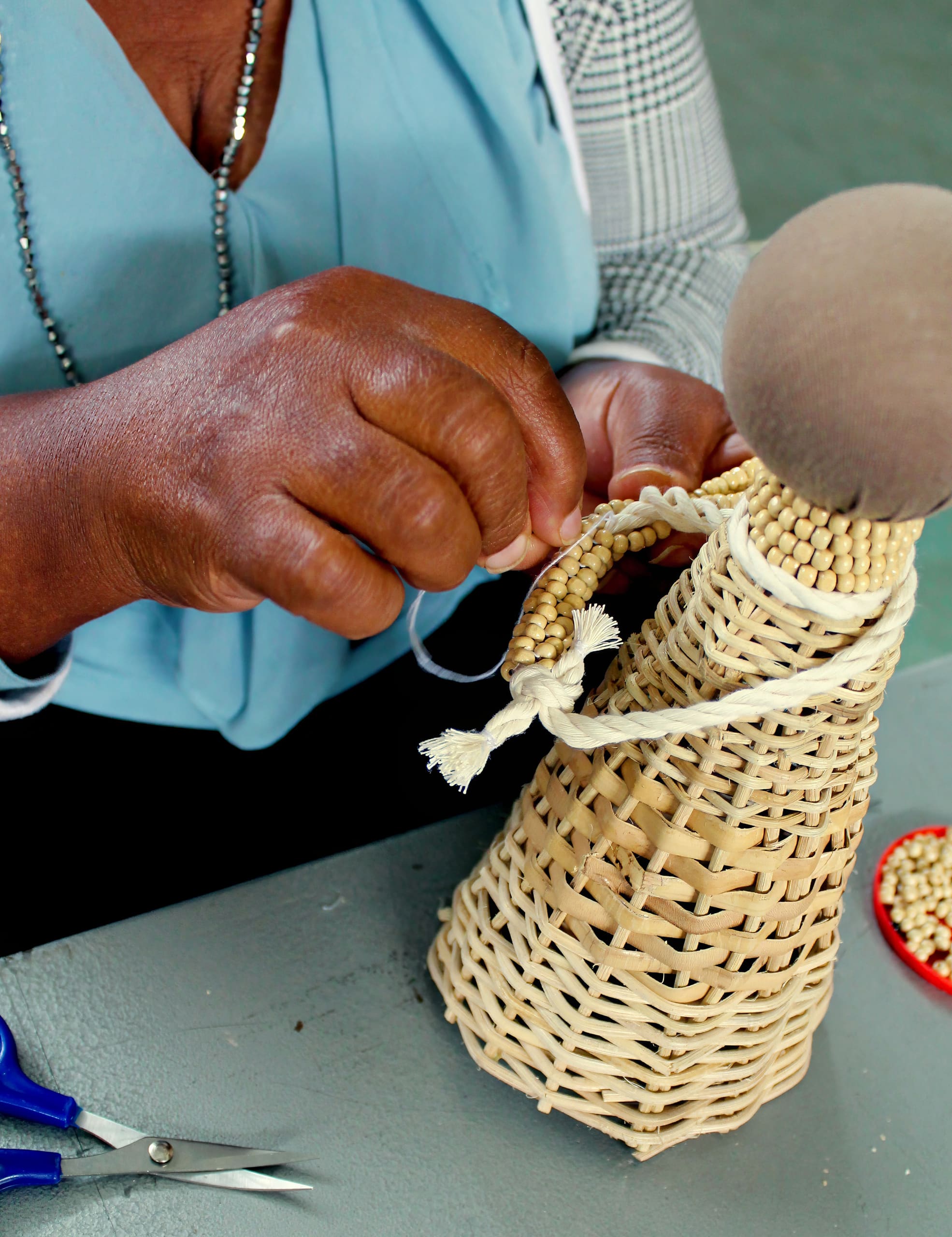 A person with dark skin threads beads onto a braided rope, attaching it to a woven basket doll with a fabric-covered head. The table holds blue-handled scissors and loose beads. The person wears a blue top and a checked jacket.