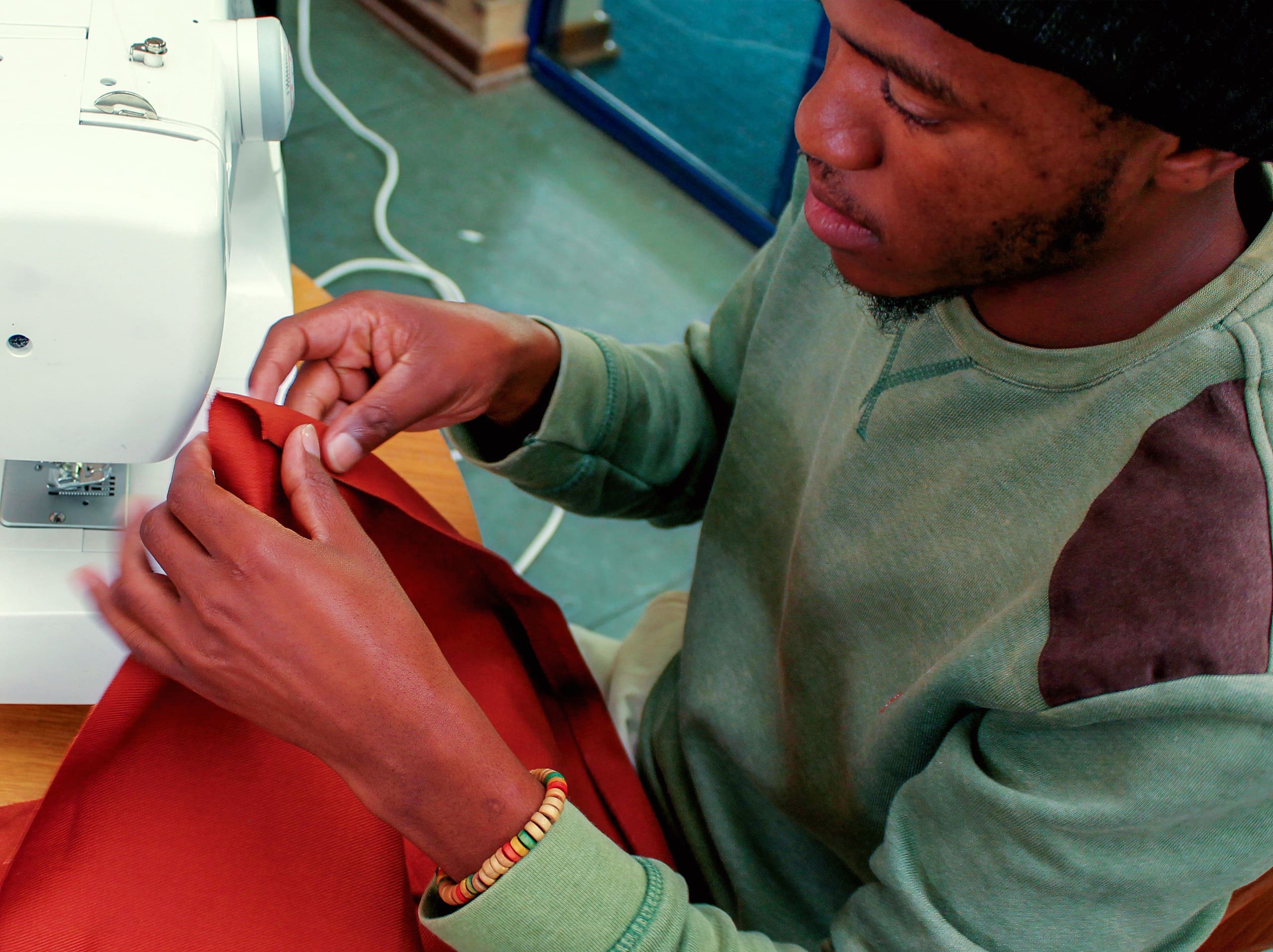 A person in a green jumper and black hat carefully guides reddish-brown fabric through a white sewing machine, focusing intently on their work. Their hands adjust the fabric, and a beaded bracelet is visible on one wrist.