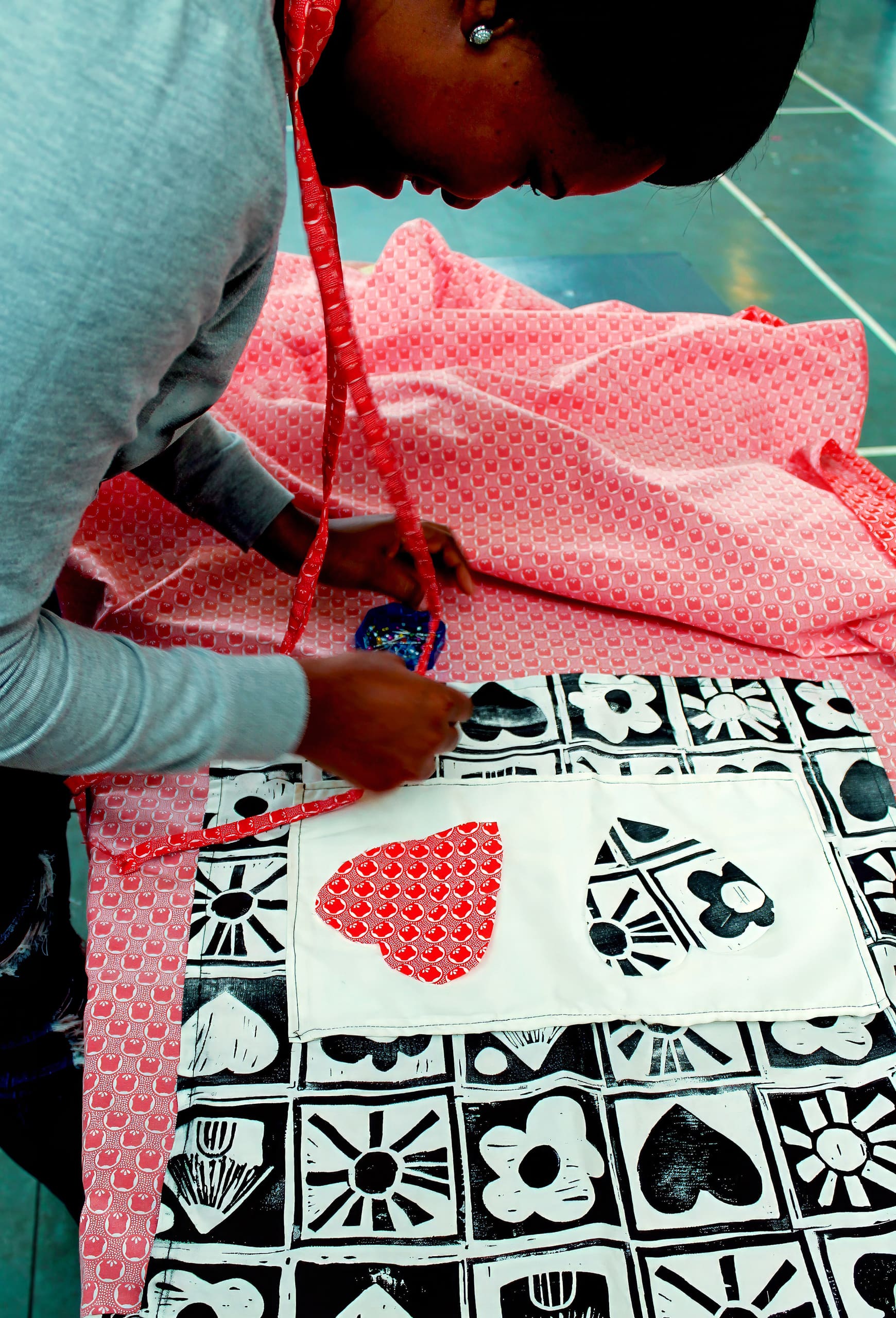 A person leans over a pink patterned fabric, working on a black and white cloth with flower and heart prints. They hold a pin cushion and a pencil, with a red measuring tape draped around their neck, focused on their sewing project.