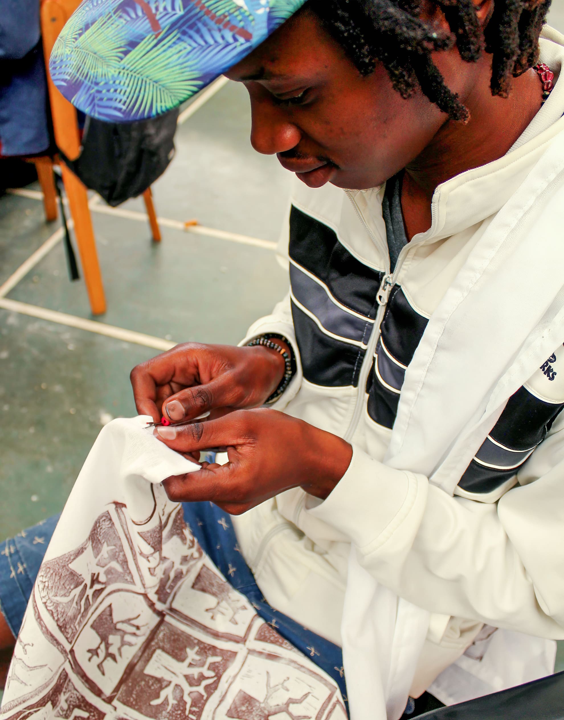 A person wearing a white jacket and a colourful cap is sitting and carefully sewing a white cloth with a brown animal and tree print. Their head is tilted down and hands are focused on stitching, highlighting attention to detail and craftsmanship.