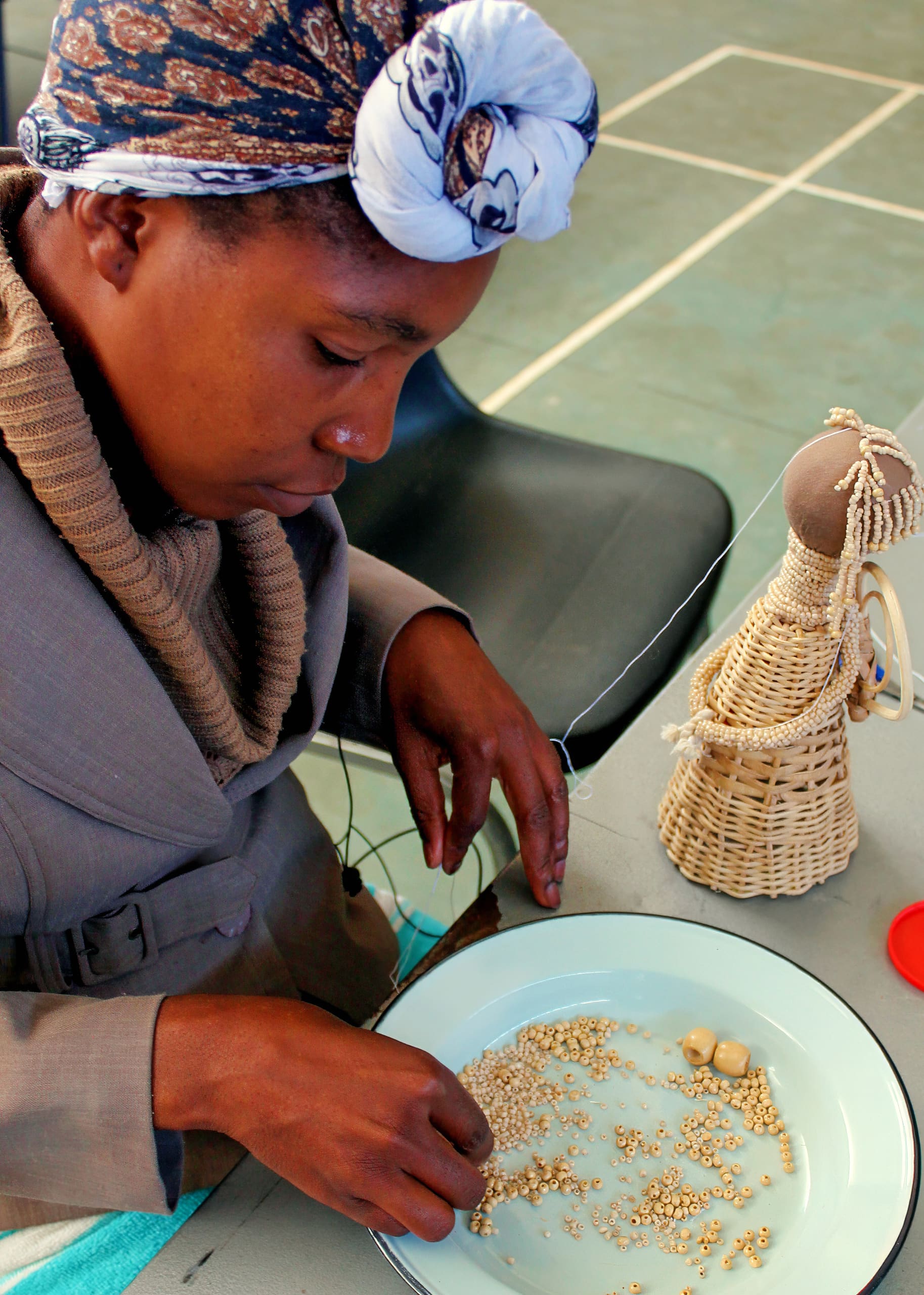 A woman in a patterned headscarf and brown jacket sits at a table, sorting small beads on a plate. Beside her is a woven doll. She appears focused, using her hands to separate beads. The background includes a black chair and tiled floor.