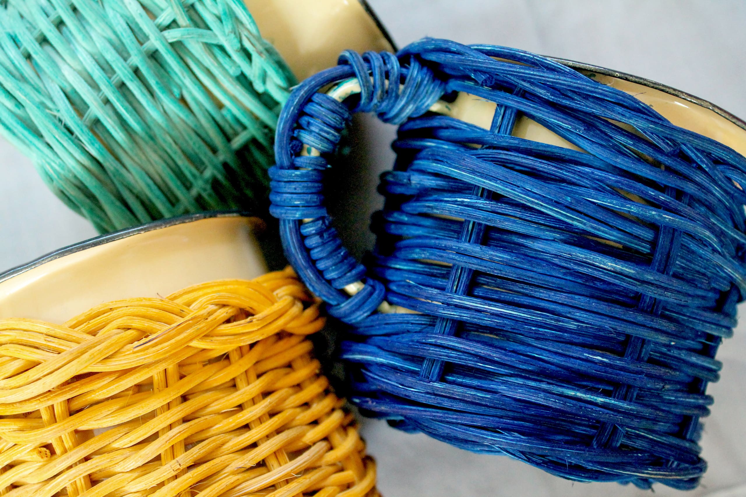 Close-up of three ceramic mugs wrapped in colourful woven materials: one mug with turquoise, another with blue, and another with yellow. The mugs are overlapping and positioned diagonally on a light surface.