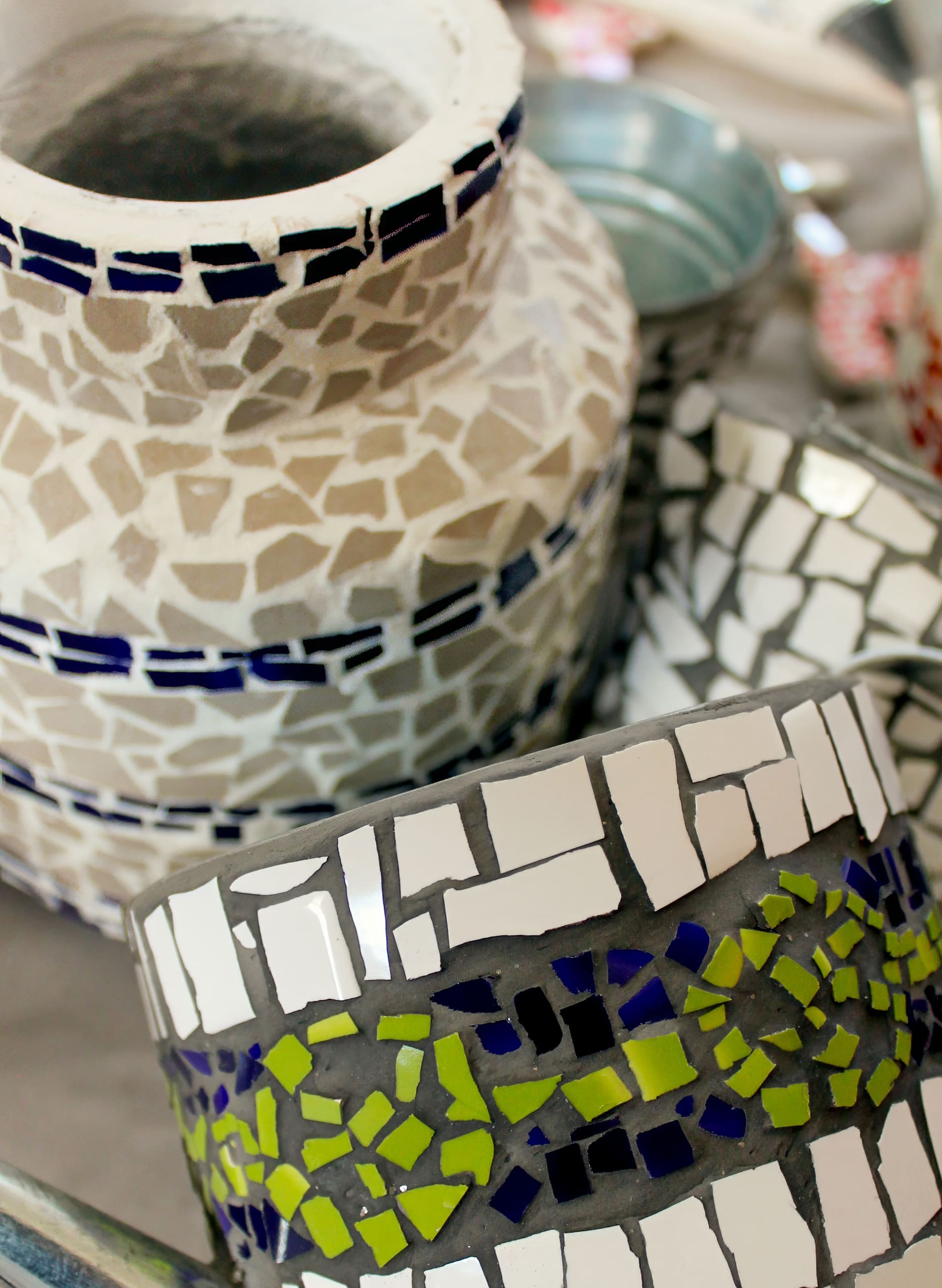A close-up of a large vase and a bowl decorated with mosaic tiles. The vase has beige and white pieces with blue bands, whilst the bowl features white, black, and green tiles in a geometric pattern. Other blurred containers are visible in the background.