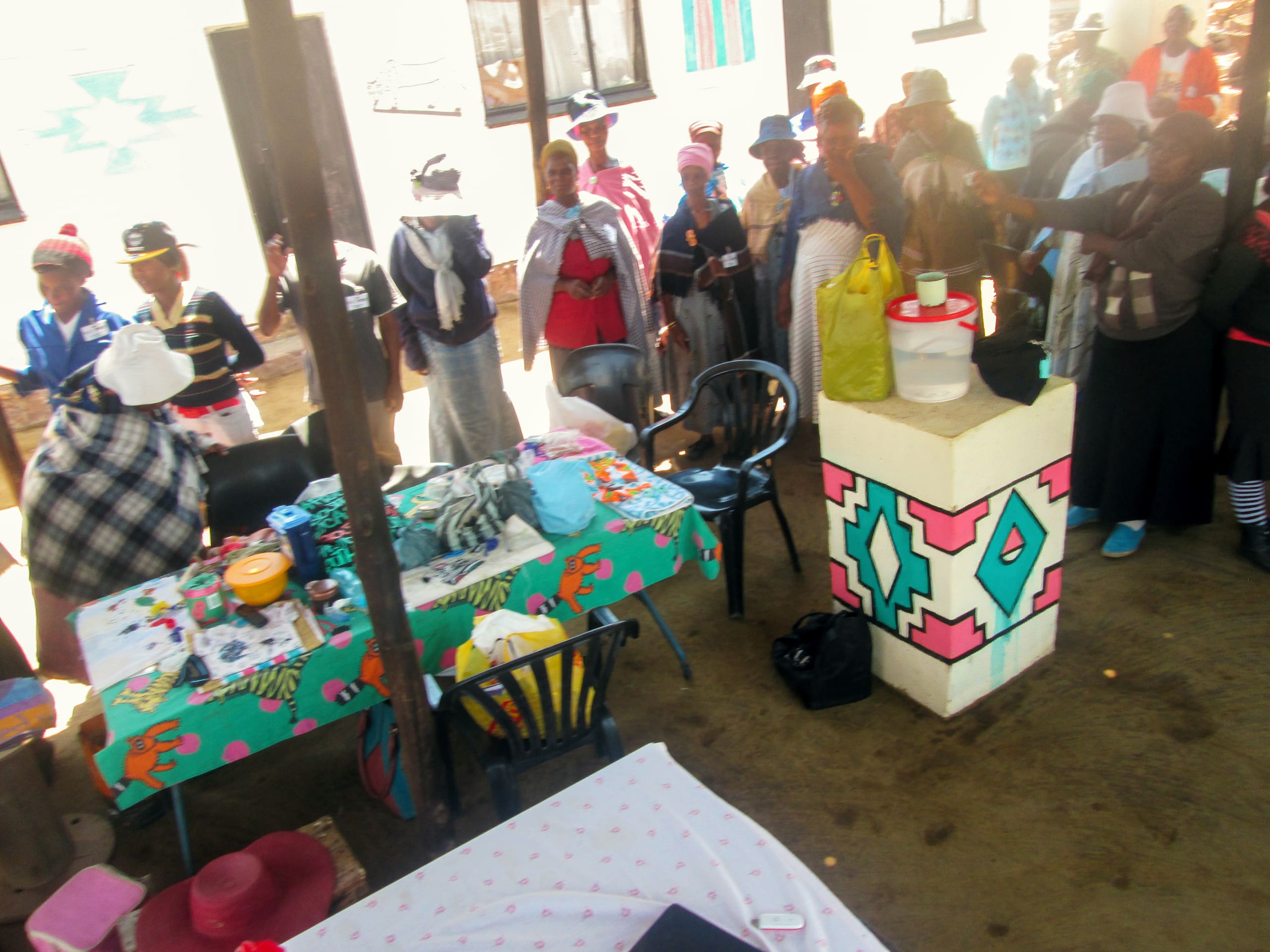 A group of people, mostly women in hats and shawls, stand in a queue indoors near a table covered with colourful cloth and supplies. The room is decorated with painted patterns on the walls and pillar. Some plastic chairs and bags are also visible.