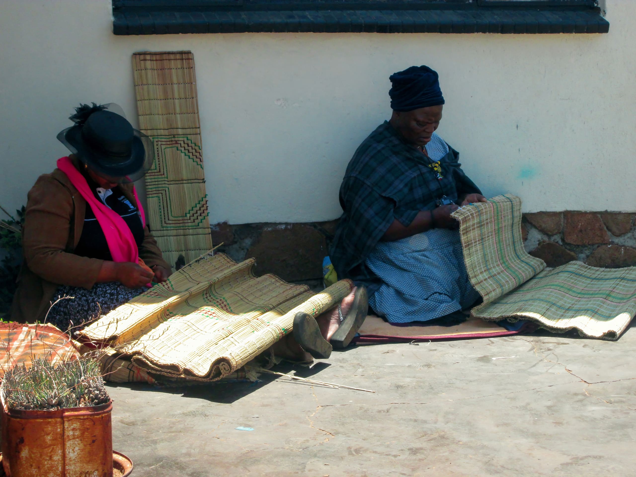 Two women sit on the ground outside, weaving traditional mats from straw. One wears a hat and pink scarf, the other a blue headscarf and checked shawl. Rolled and finished mats are beside them, with a small potted plant in the lower left corner.