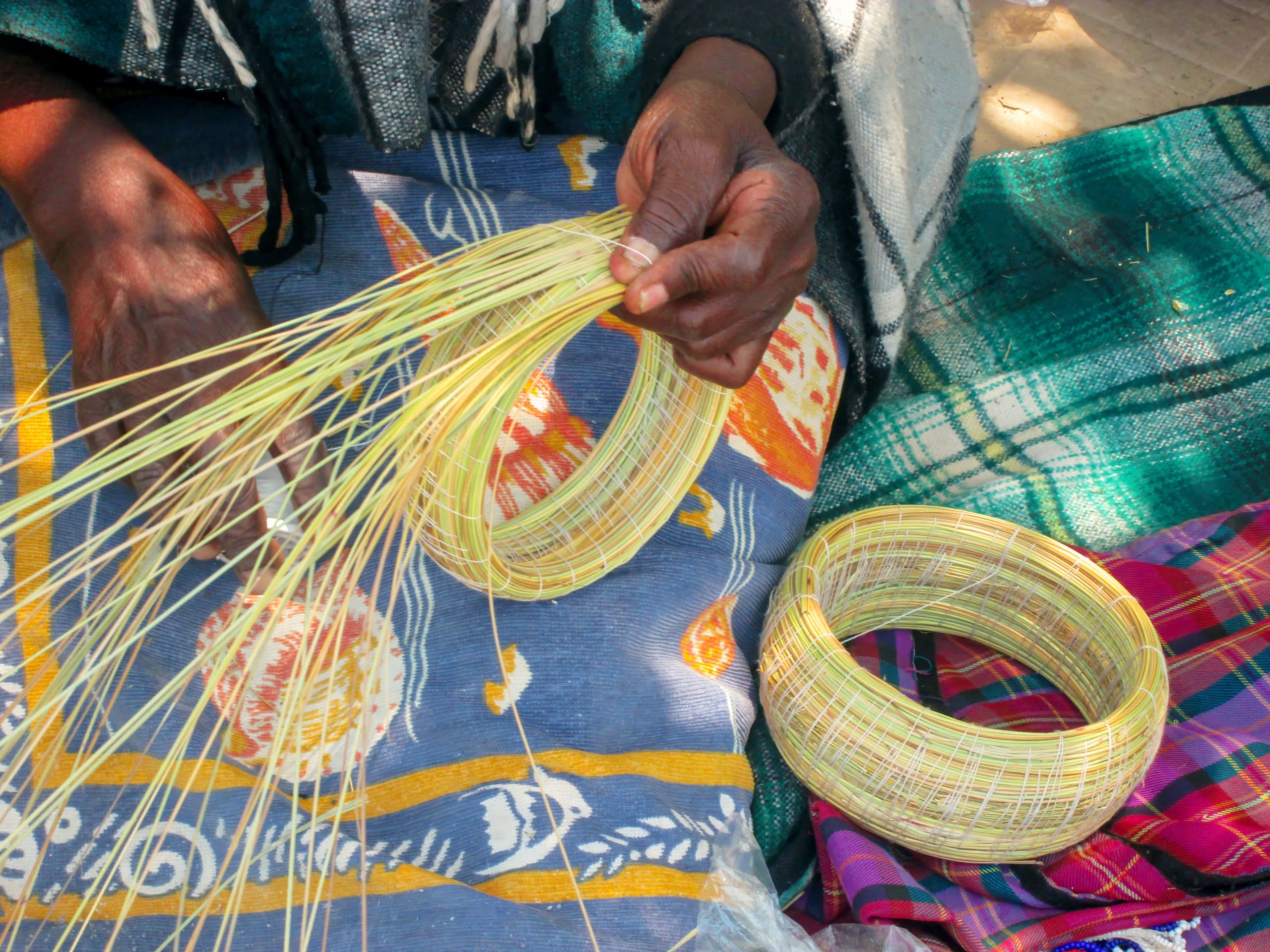 Close-up of hands weaving thin, yellow palm fibres into circular baskets. The person works over a blue cloth with orange and white patterns, and nearby are finished baskets. A green and purple checked cloth is also visible in the background.