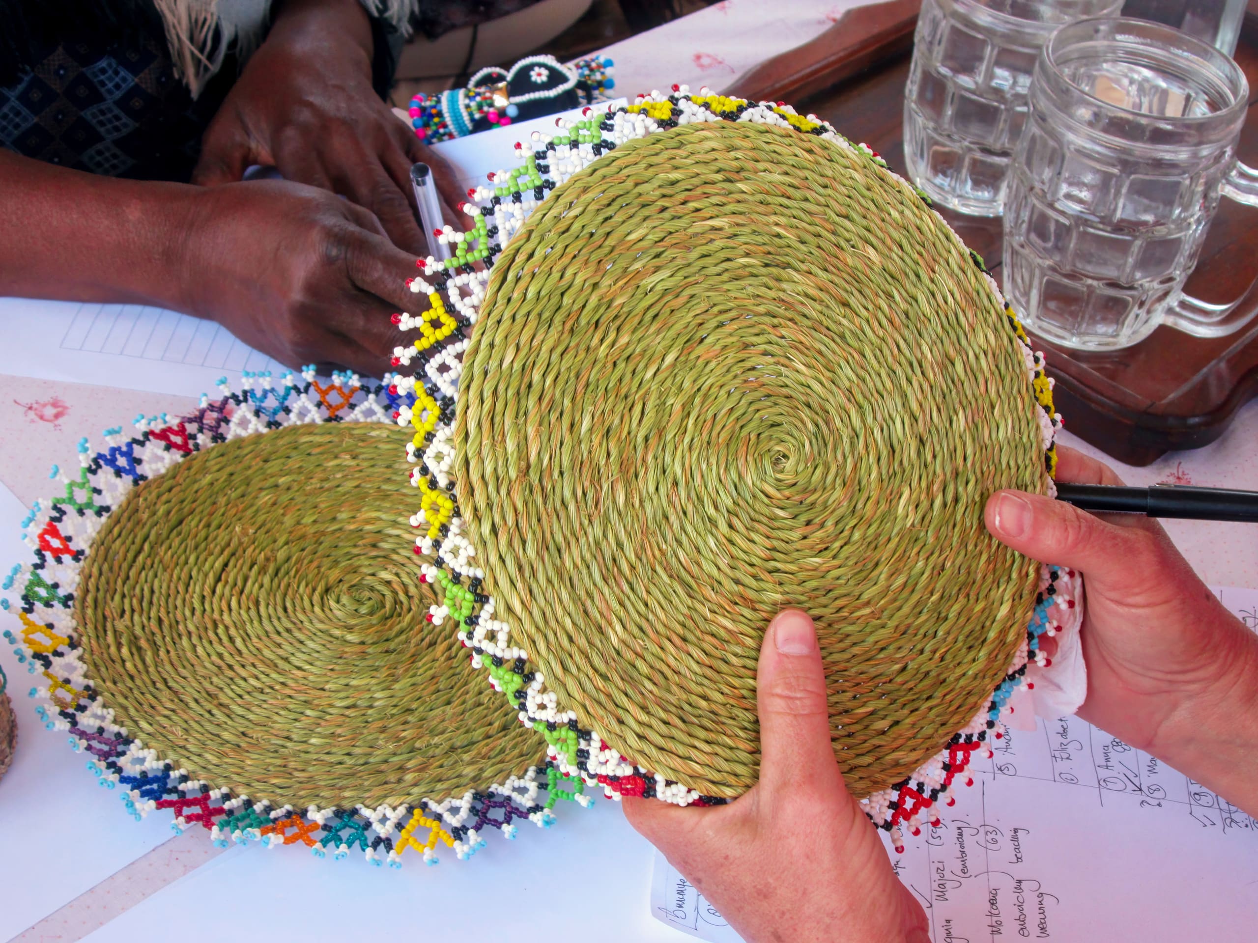 Two people hold round, woven grass mats decorated with colourful beadwork around the edges. The mats rest on a table with papers, pens, and glass mugs. One person has lighter skin, the other darker; only their hands and part of their arms are visible.
