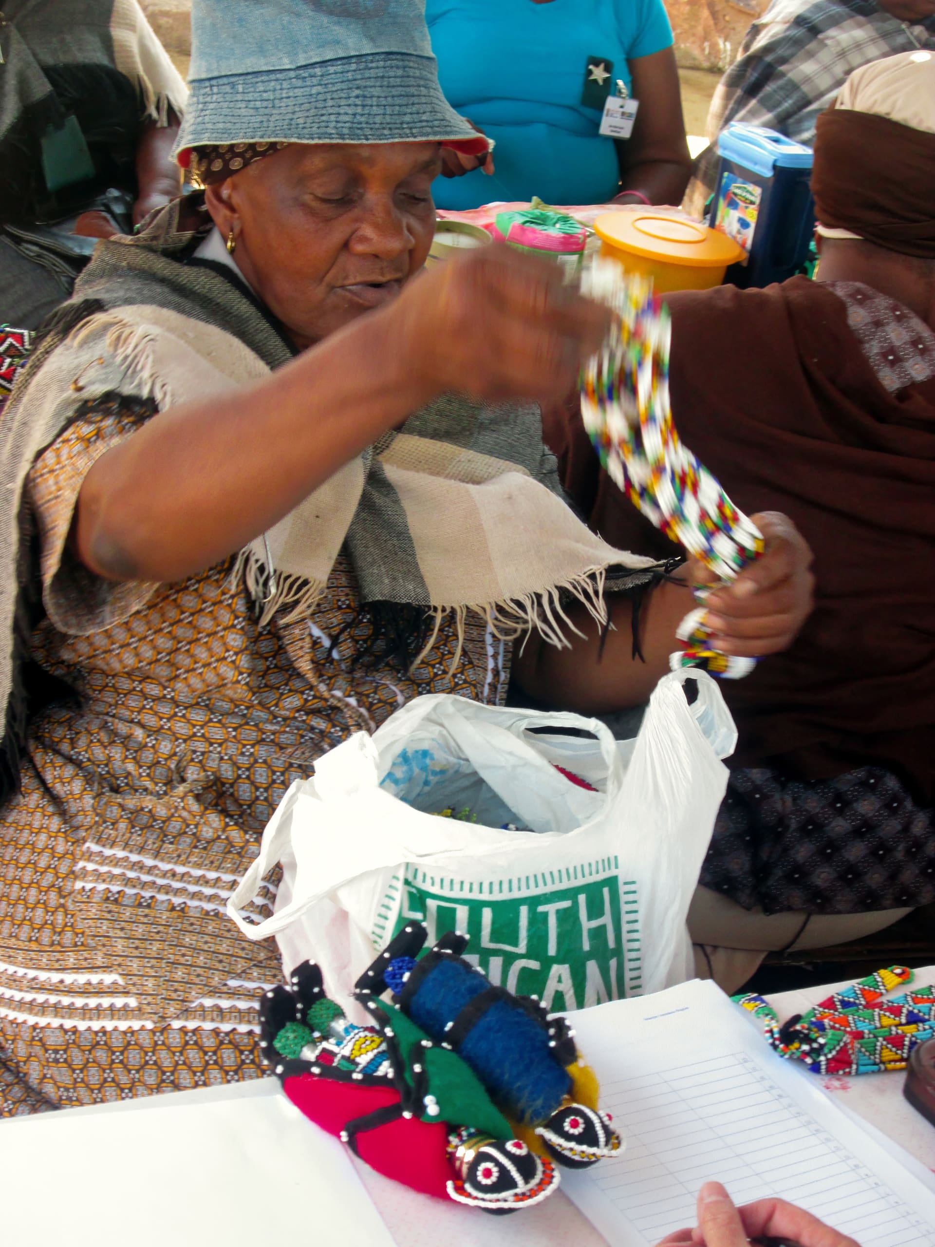 An elderly woman in a patterned dress and hat holds colourful beaded crafts at a table. In front of her is a white bag labelled “SOUTH AFRICAN,” beaded items, and a clipboard with paper. Other people and containers are partially visible in the background.