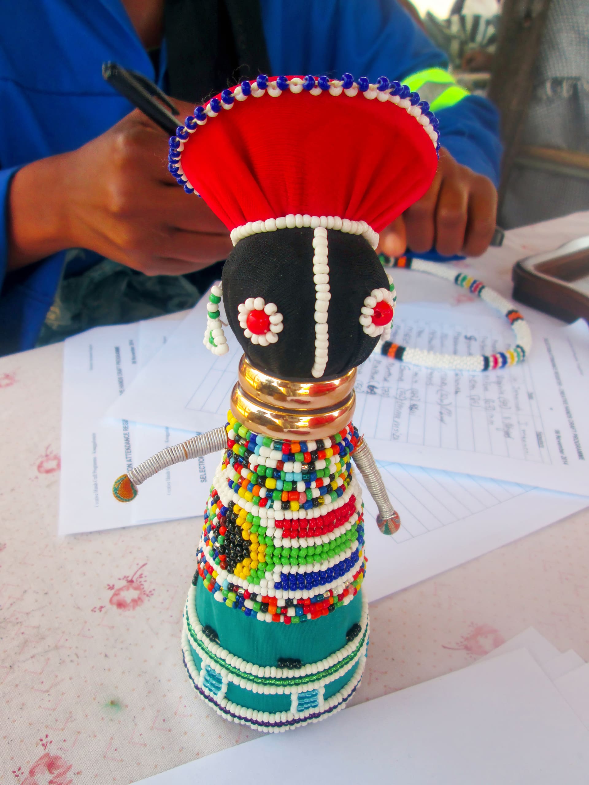 A colourful beaded doll with a red headdress, gold rings for a neck, and a multicoloured beaded dress sits on a table. A person in blue writes on papers in the background, partially visible, with a beaded bracelet on their wrist.