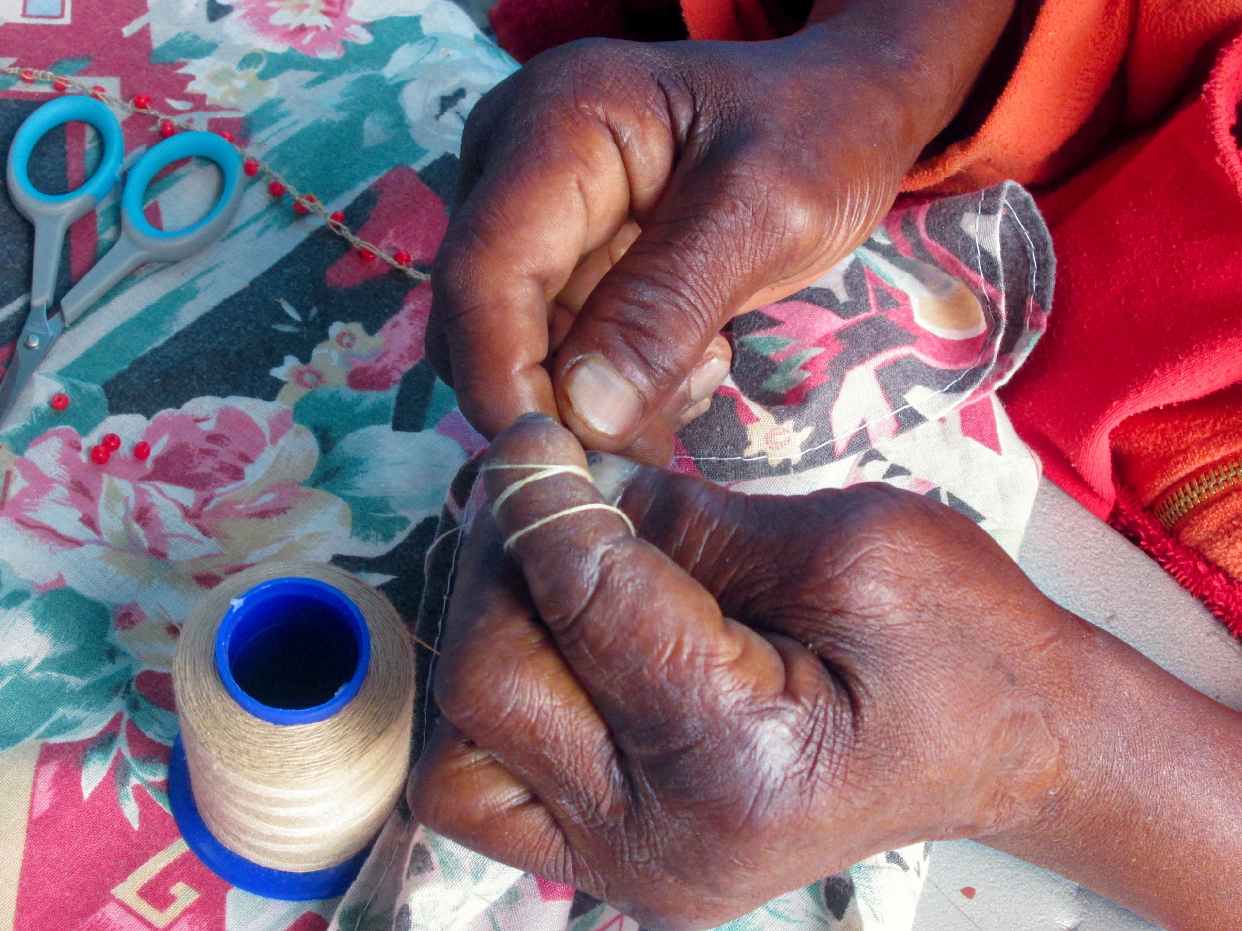 A close-up of two hands threading a needle with beige thread. The hands rest on colourful floral fabric. Nearby are a blue spool of thread and a pair of blue-handled scissors. The person wears an orange garment.