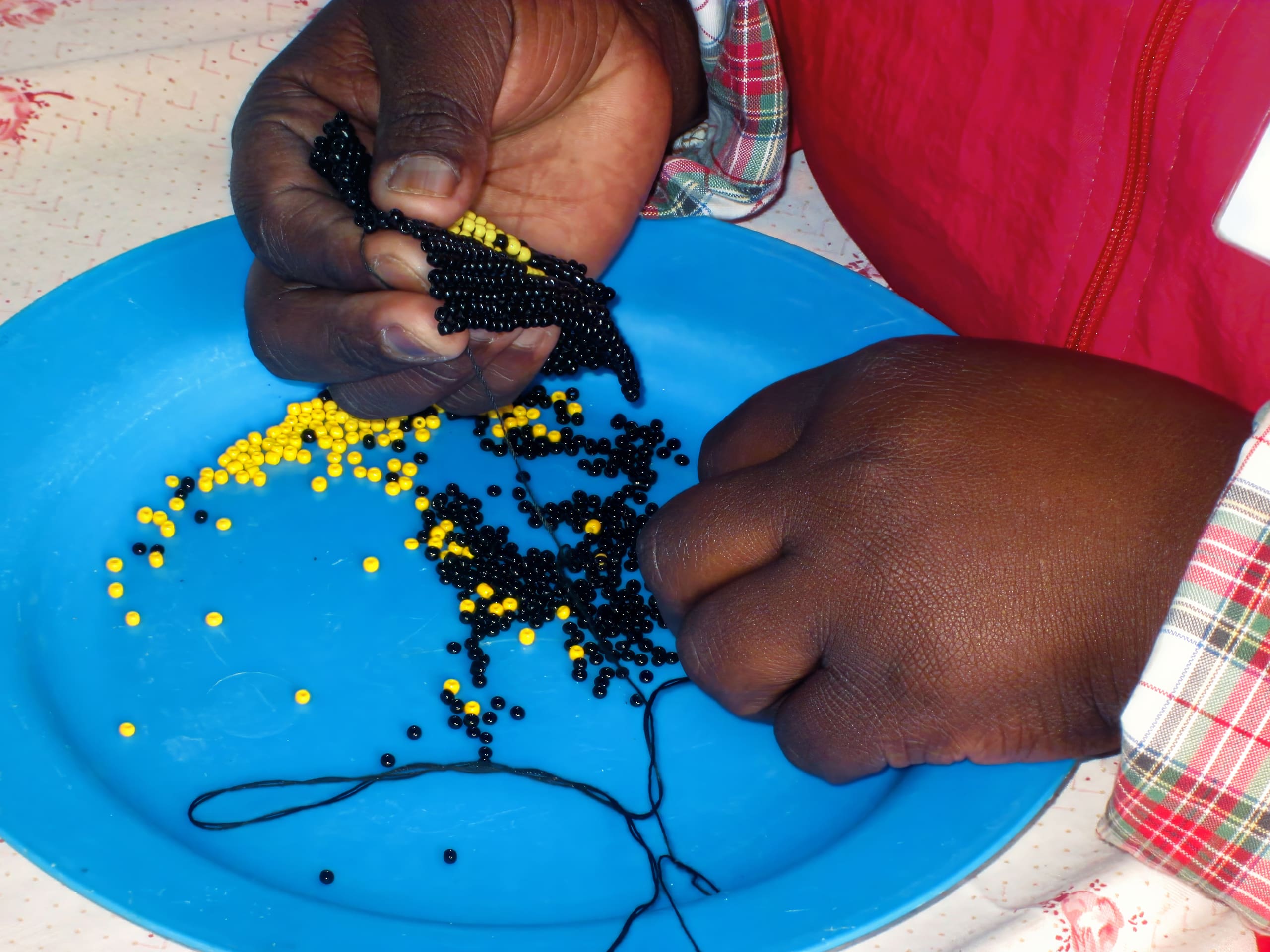 A person with dark skin threads small black and yellow beads onto black string over a blue plate. Their hands are close together, focused on the detailed work. They wear a red jacket and a checked shirt with visible cuffs.