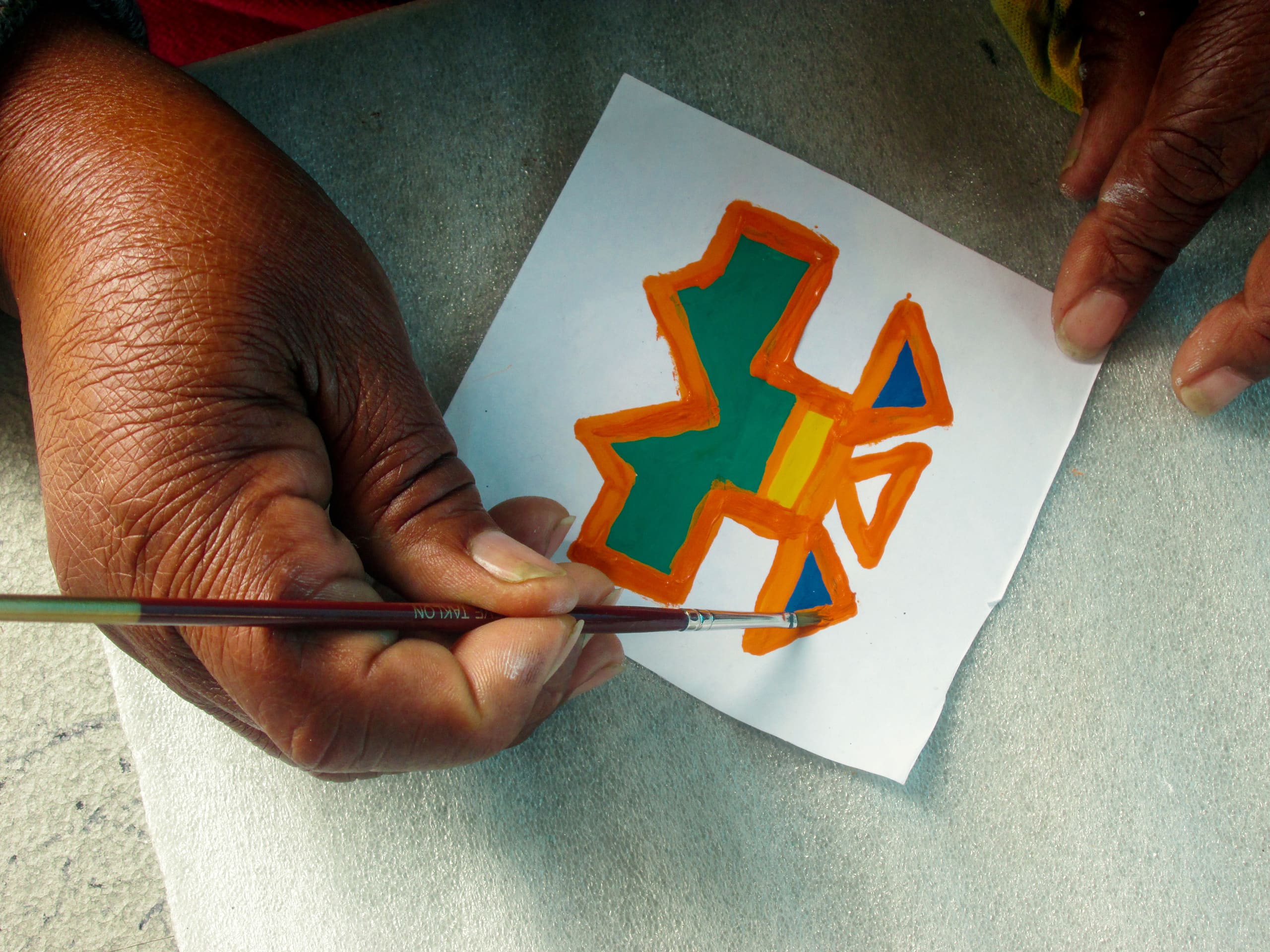 A person with medium-brown skin paints a geometric, colourful design on a small white paper. The design features orange, green, and yellow shapes. The person holds a paintbrush in one hand and steadies the paper with the other. A grey surface is underneath.