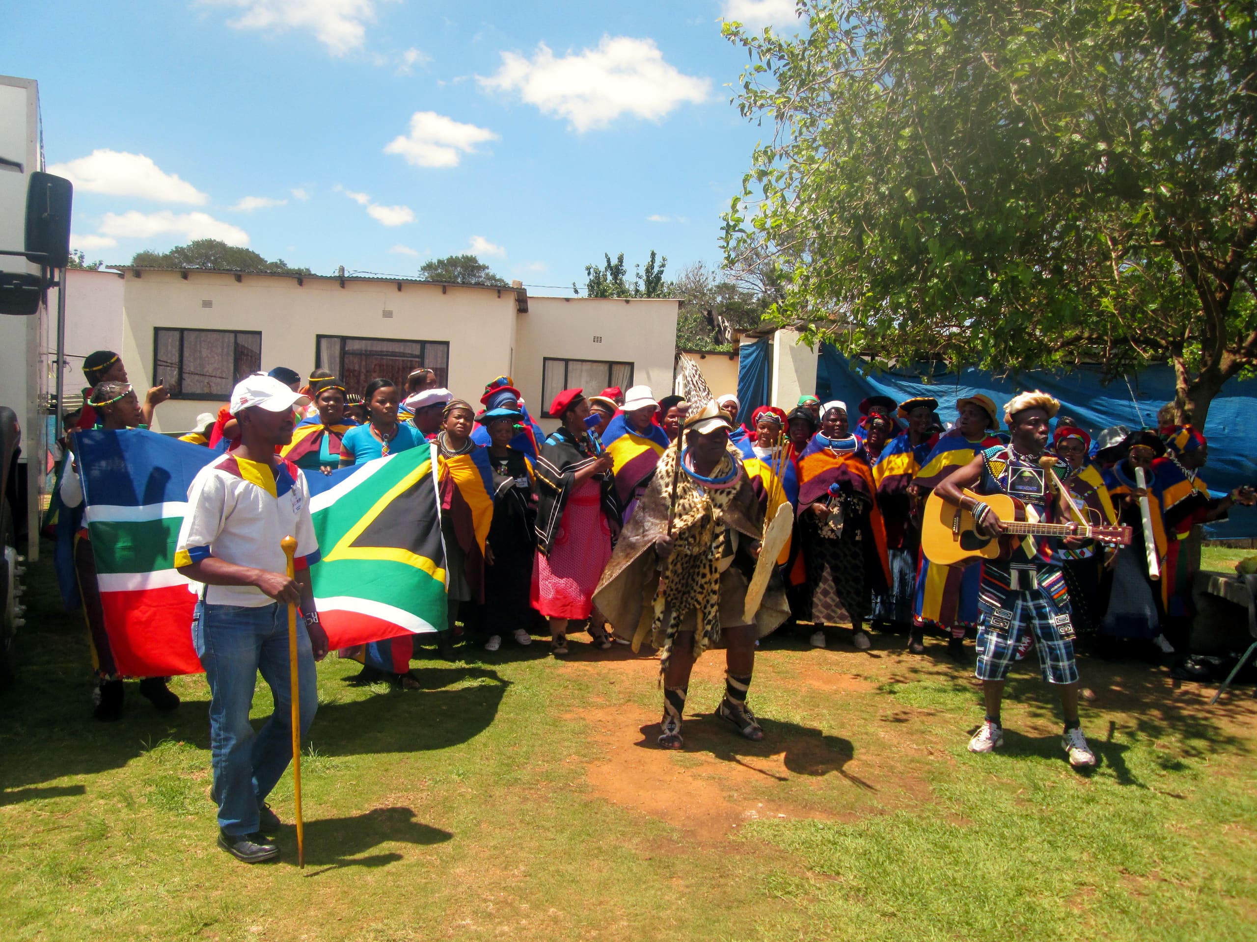 A group of people in colourful traditional attire stand outdoors, some holding South African and Namibian flags. In front, a man wears traditional clothes and another plays guitar. Trees, a building, and a blue sky are visible in the background.