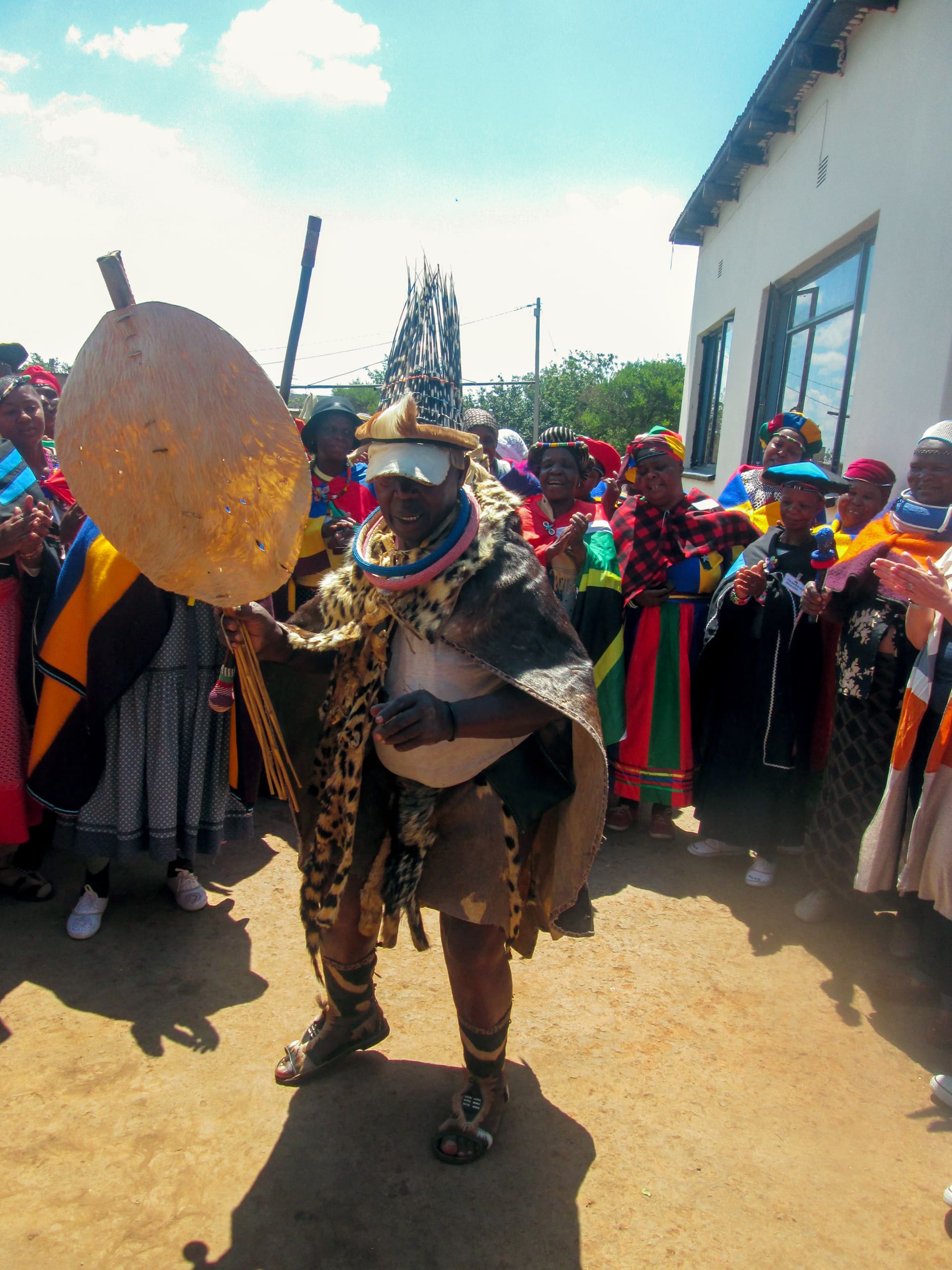 A person in traditional attire, including animal skins, bead jewellery, and a feathered headdress, dances whilst holding a shield and a stick. A group of vibrantly dressed people stand in a semi-circle, watching and celebrating outdoors near a building.