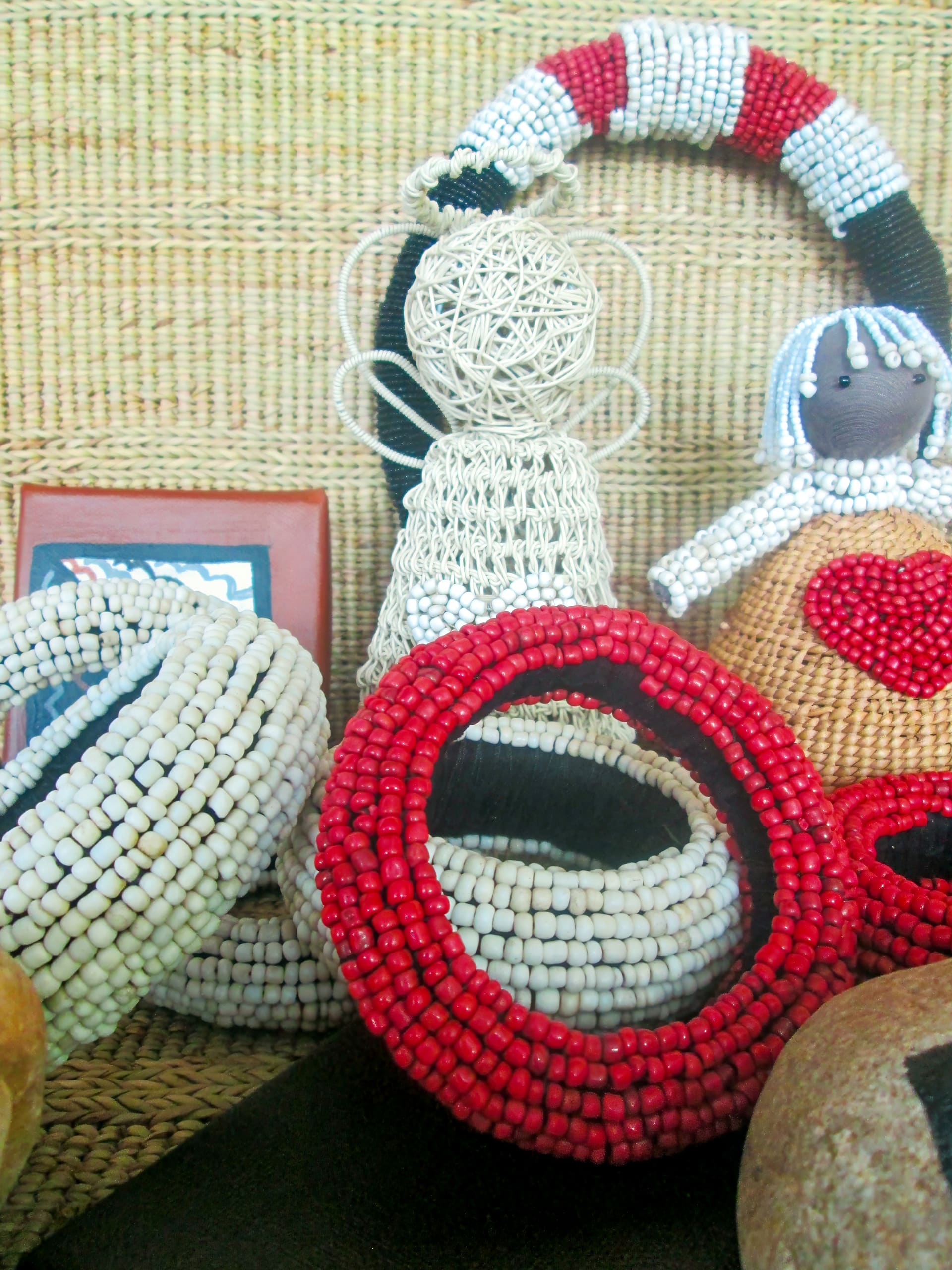 Handcrafted beadwork including white and red bracelets, a wire angel, and a doll with a stitched red heart, are displayed against a woven background. The items are arranged closely together, showcasing their textures and intricate details.