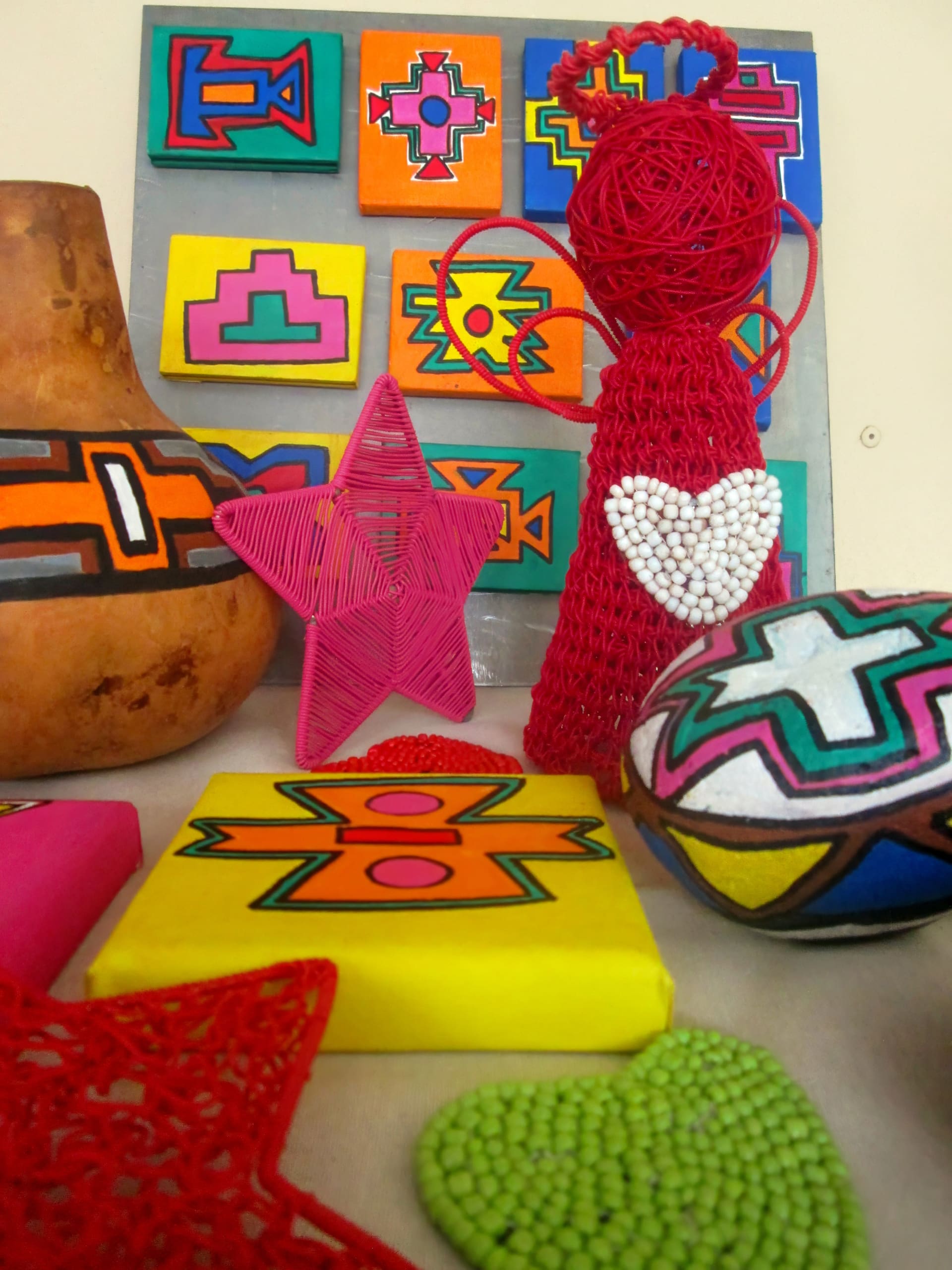 Colourful crafts on display, including a red wire angel with a beaded heart, geometric-patterned painted boxes and gourds, a pink string star, and bright green and red woven decorations, all arranged before a backdrop of vibrant, abstract artwork.