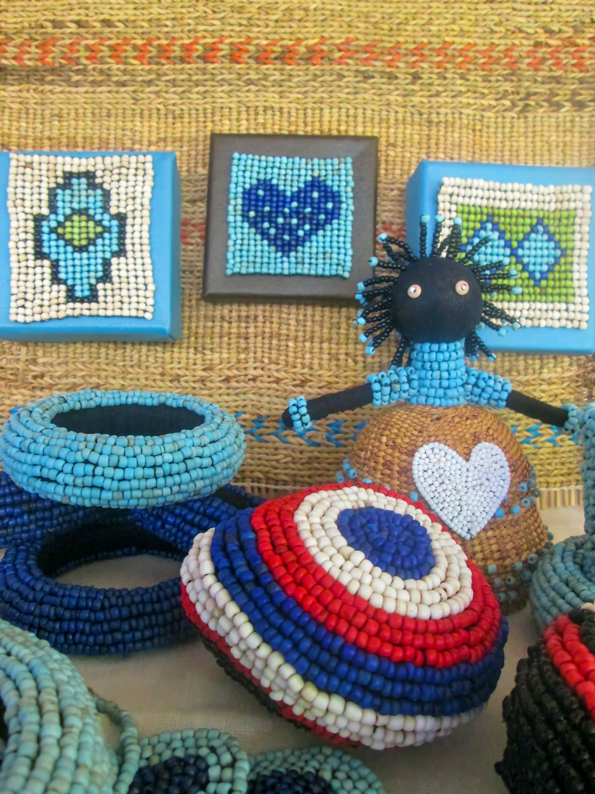 Colourful beaded jewellery including blue bangles and red, white, and blue round items are displayed with a small black doll wearing a beaded outfit. Behind them are framed, beaded geometric designs with hearts and diamond shapes in blue and green tones.