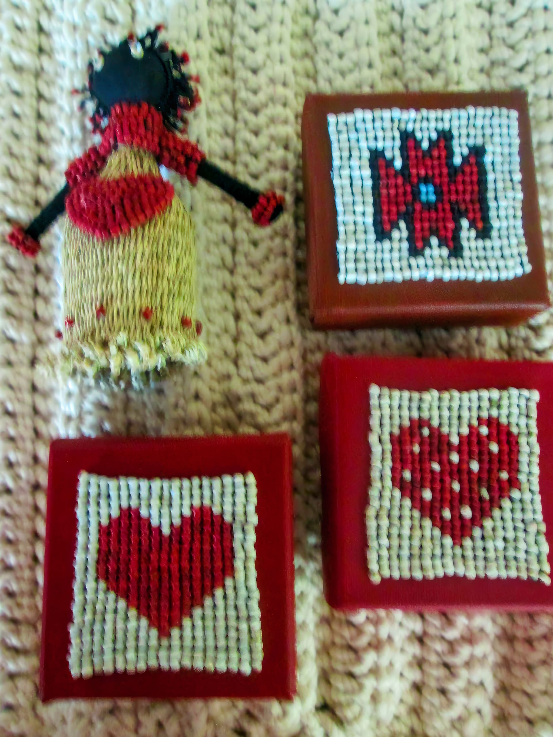 A handmade doll with a straw skirt and red scarf lies on a knitted beige surface beside three small red boxes decorated with white beaded designs: a flower, a heart, and a heart with polka dots.