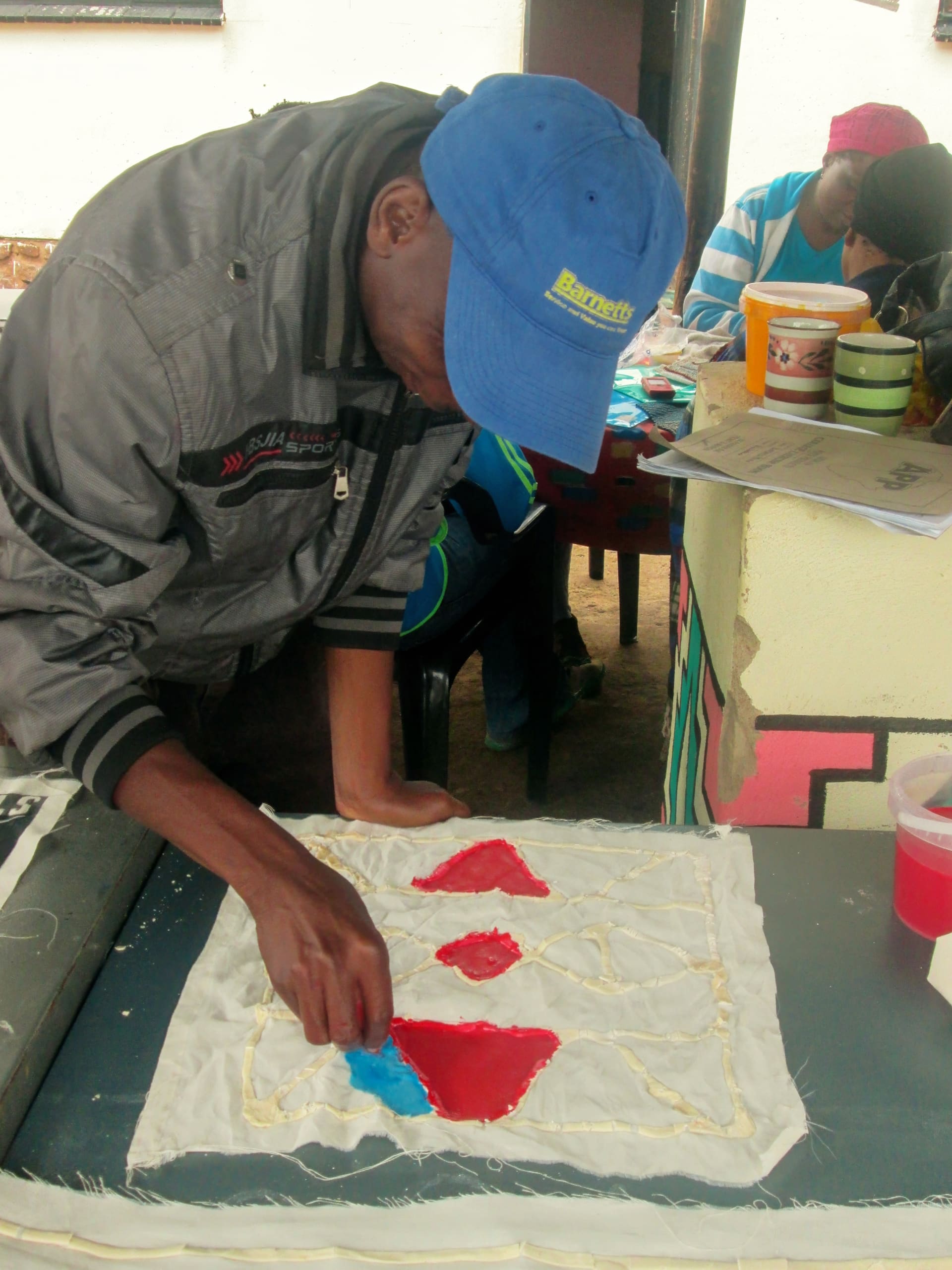 A person in a blue cap and grey jacket bends over a table, painting blue dye onto white fabric with raised wax outlines and red shapes. Other people and art supplies are visible in the background in a creative indoor setting.