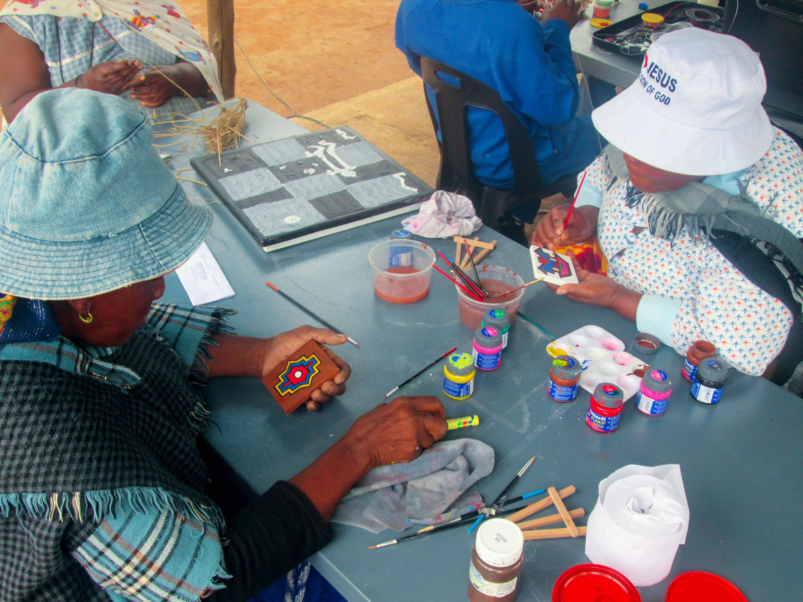 Two women wearing hats and traditional clothing sit at a table painting small wooden crafts with colourful paints. Art supplies, paint jars, and brushes are scattered on the tabletop. Another person works in the background, partially visible.