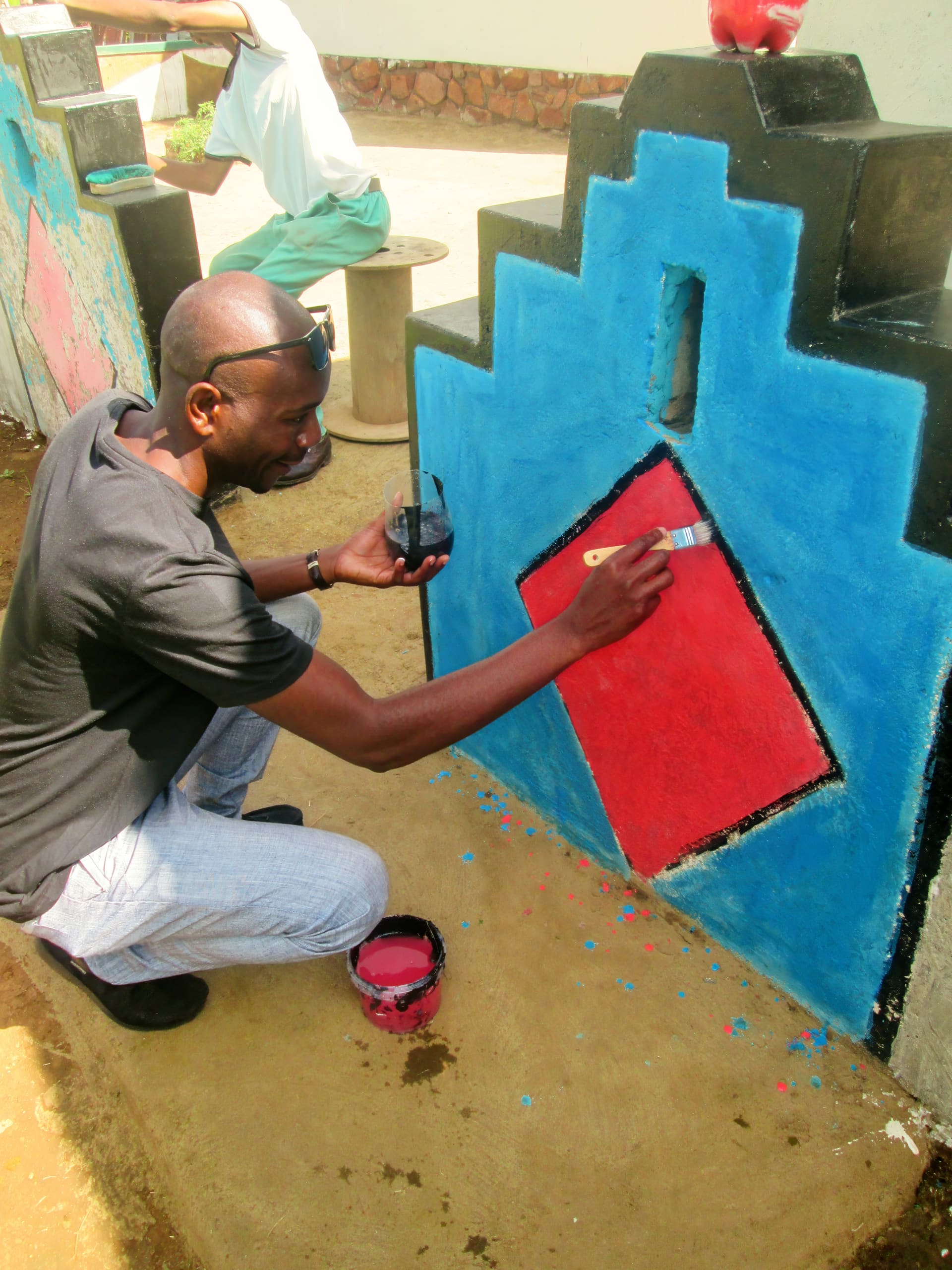 A man in sunglasses kneels outdoors, painting a geometric mural on a wall. He holds a cup of paint and uses a brush to fill in a red diamond shape bordered by blue and black. There’s another person and artwork visible in the background.