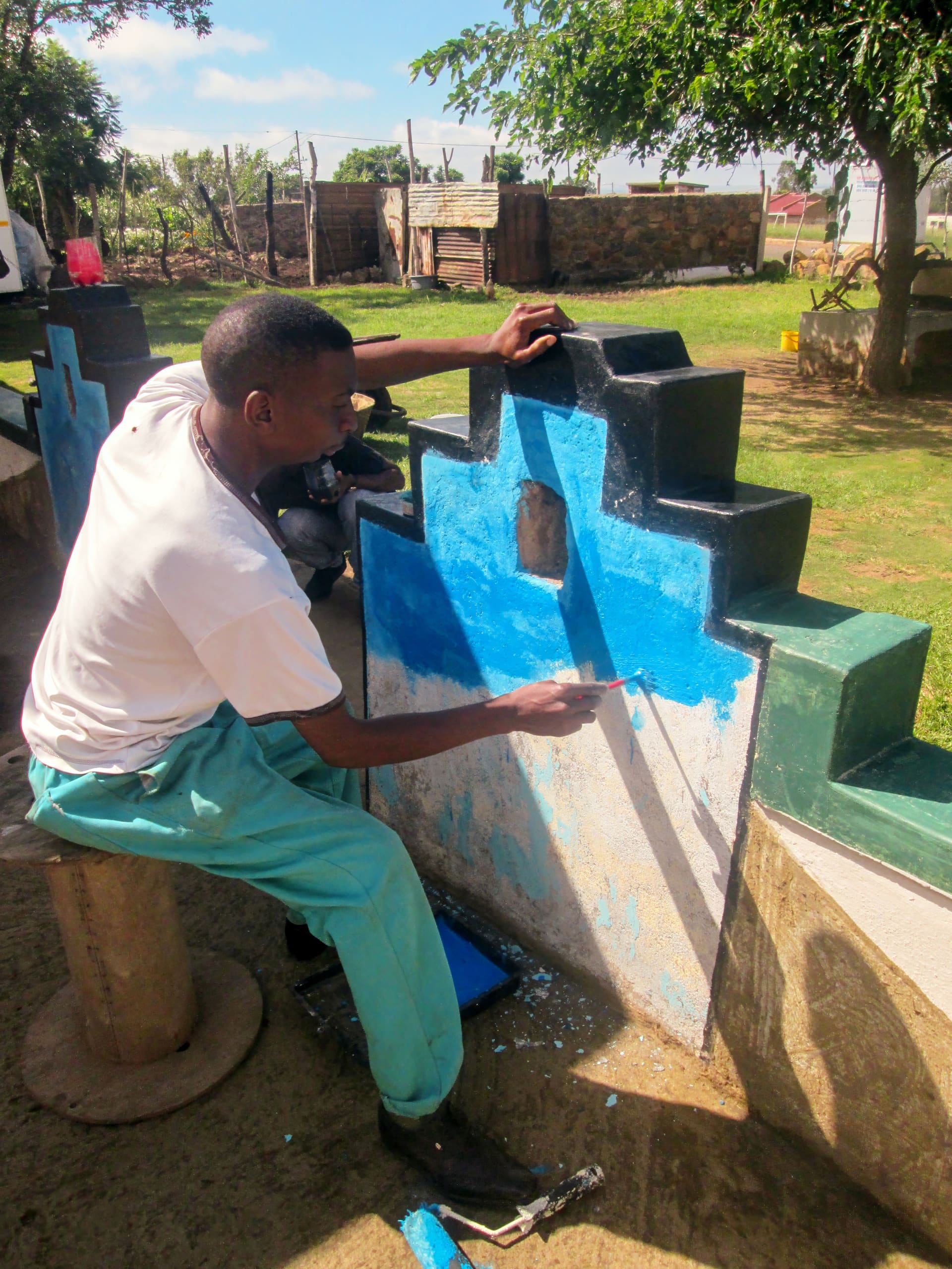 A man sits on a stool outdoors, painting a geometric concrete wall bright blue. He wears a white shirt and teal trousers and uses a small brush. Green grass, trees, and wooden fencing are visible in the sunny background.