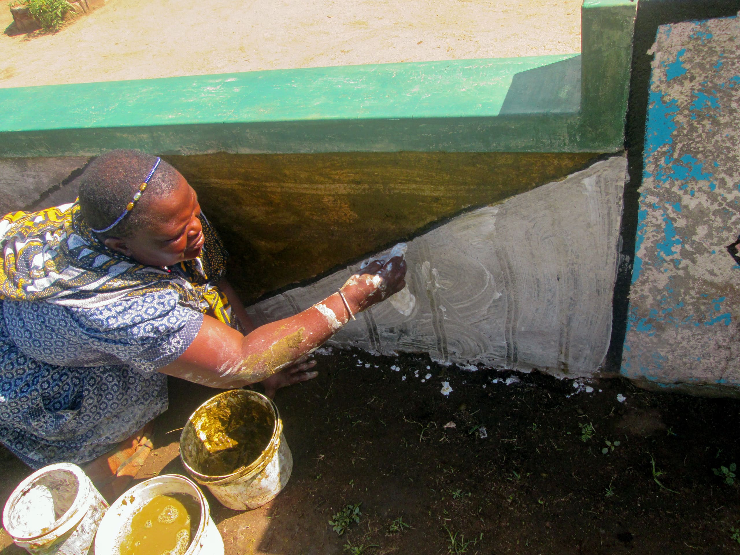 A woman kneels outside by a concrete wall, applying white paint or plaster to its lower section. She wears patterned clothing and a headband. Several paint buckets sit nearby on dark soil, and the upper part of the wall is painted green.