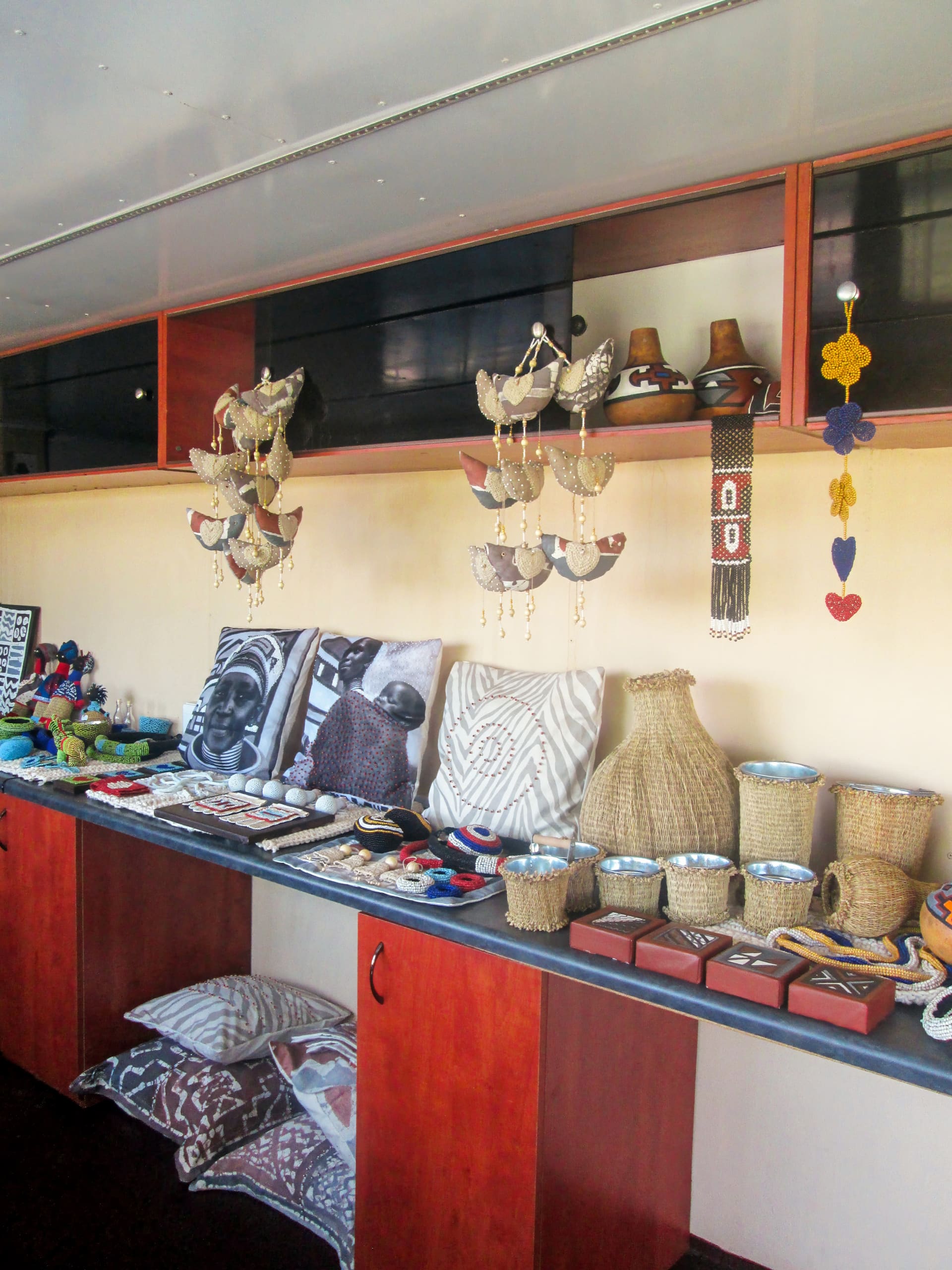 A display of handcrafted items on a worktop, including woven baskets, pottery, beaded jewellery, cushions with patterned covers, and small decorative items. Some crafts, like beaded ornaments and wall hangings, are arranged on the wall and shelves above.