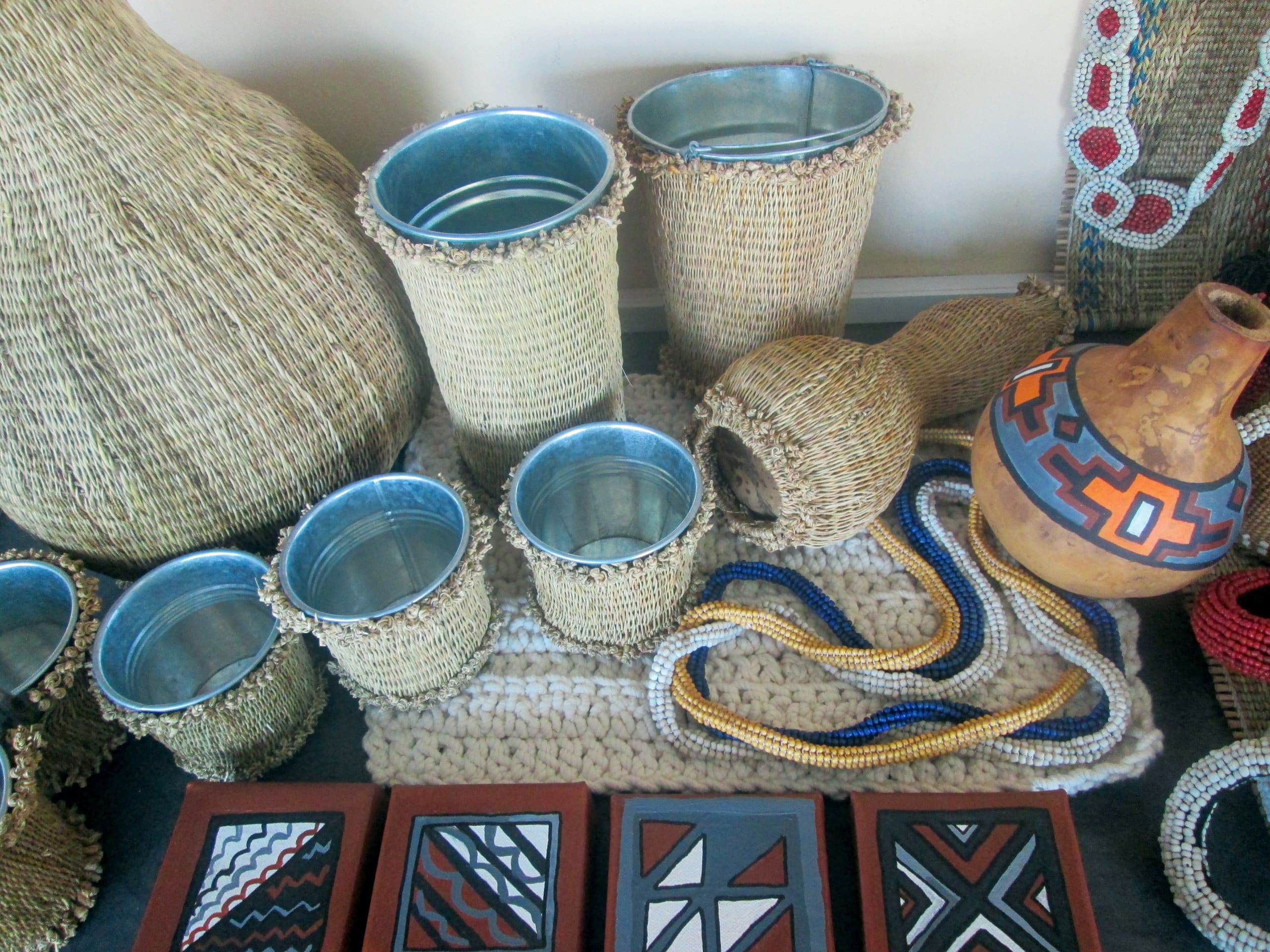 A display of traditional African crafts, including woven baskets with metal linings, a patterned clay pot, colourful beaded necklaces, a thick woven mat, and painted wooden plaques featuring geometric designs, all arranged neatly on a surface.