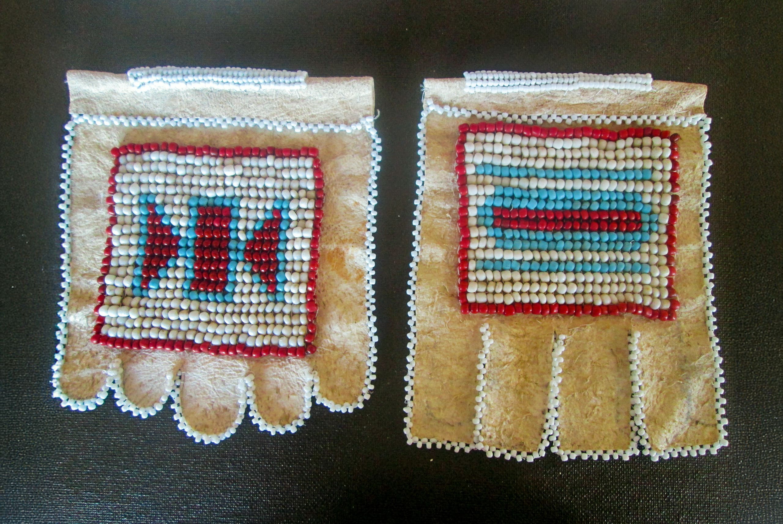 Two pieces of tan leather, shaped like partial gloves, are decorated with colourful beadwork in geometric patterns. Each has a beaded border and square designs in red, white, and blue beads on the top surface. The background is black.