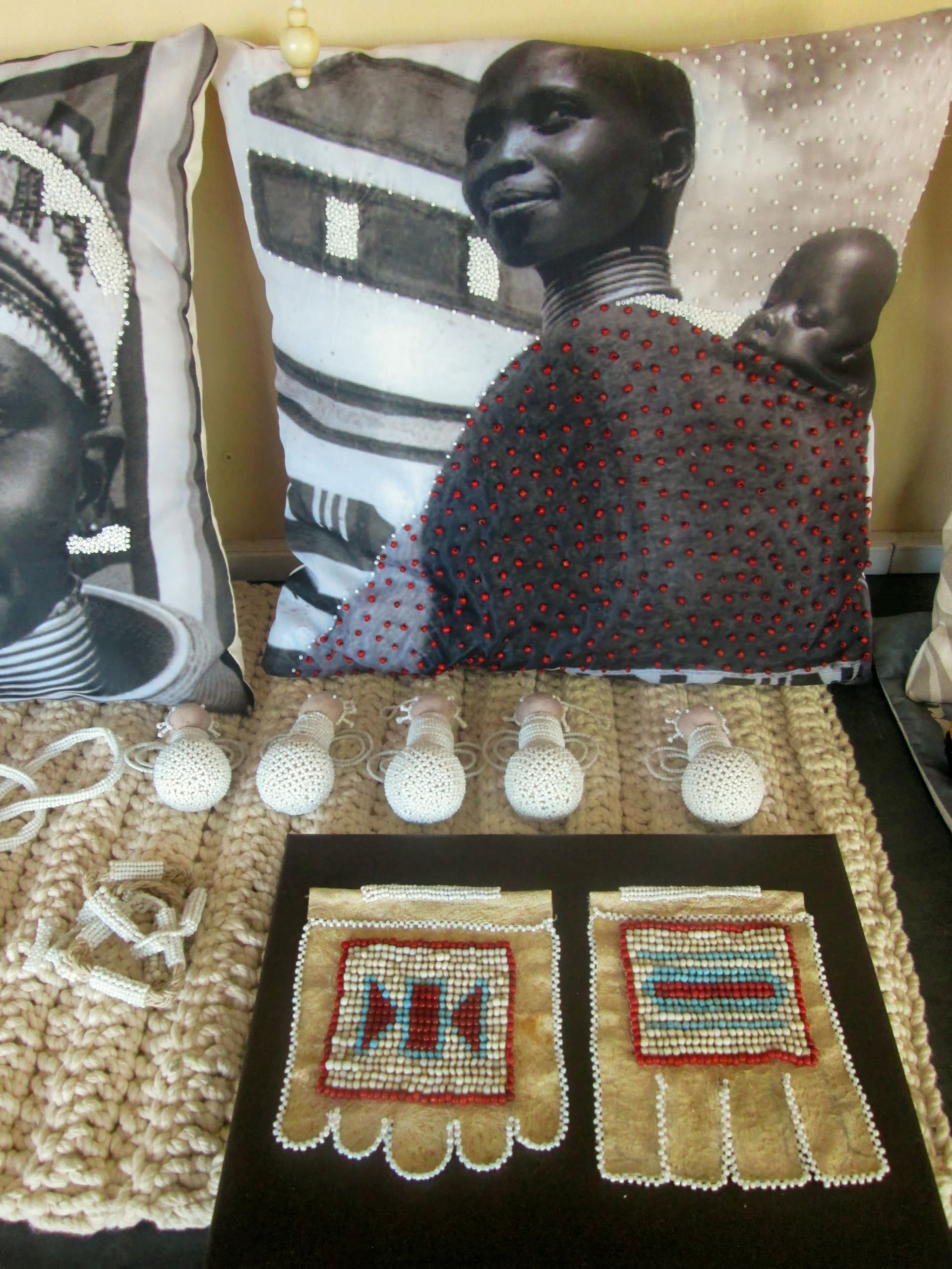 A decorative cushion features a black-and-white photo of an African woman carrying a baby, with red and white beads sewn onto the image. In front are crocheted items, beaded ornaments, and two rectangular beaded patches with geometric patterns on tan fabric.
