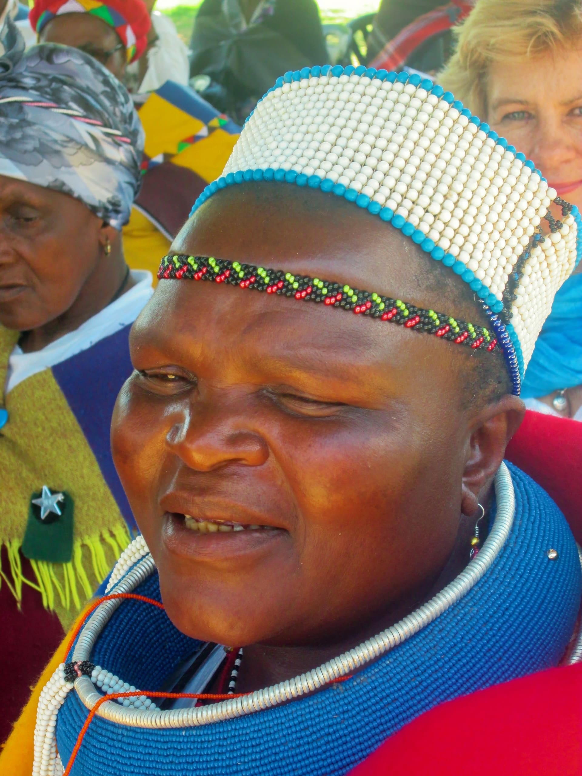 A woman wearing a beaded white headdress, colourful bead necklace, and red and blue clothing smiles slightly. Other women in vibrant attire and headscarves are in the background, with one woman in a blue scarf and another wearing yellow and green.