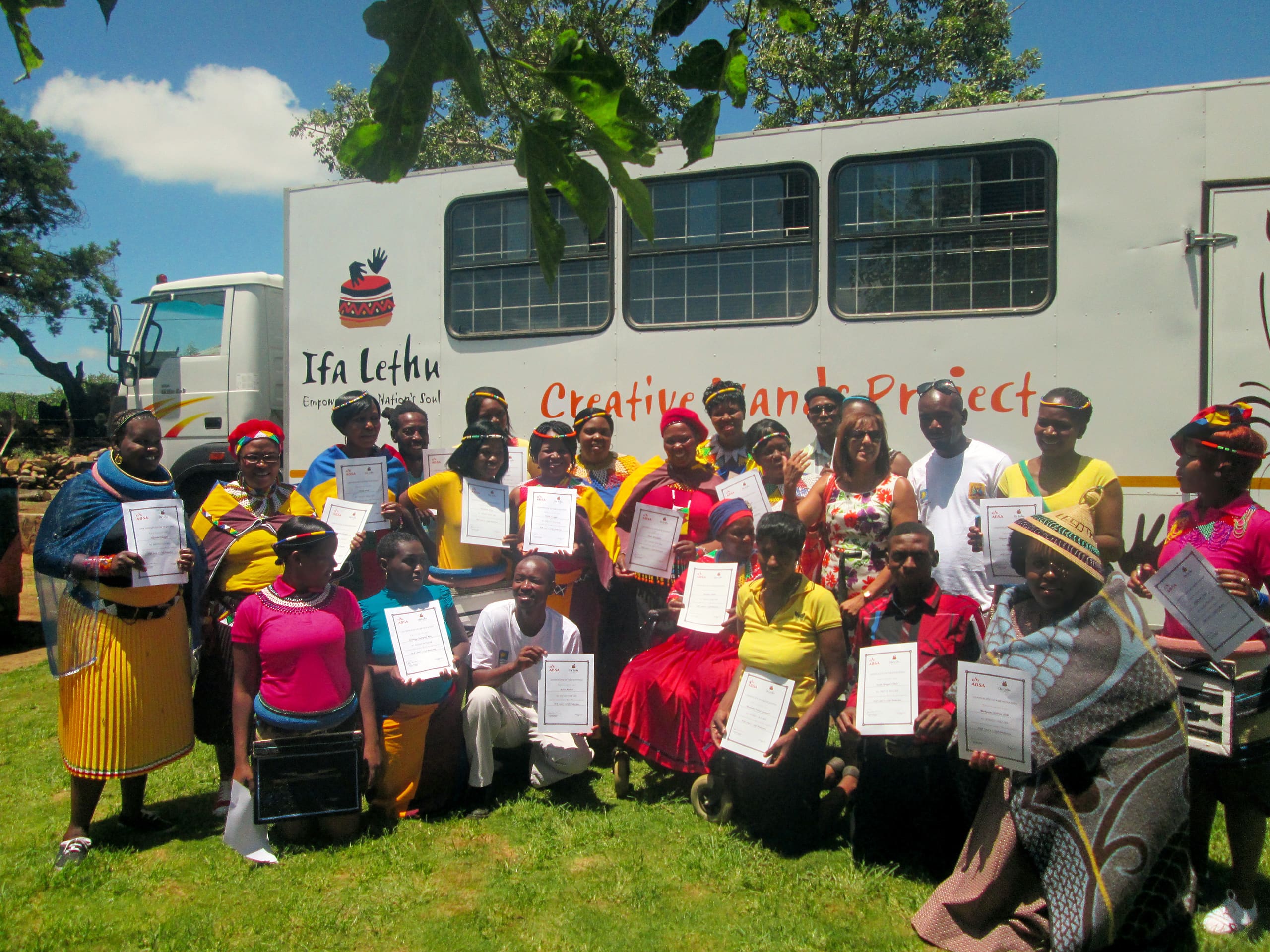 A group of people, mostly women in colourful traditional attire, pose outdoors in front of a large white lorry with 