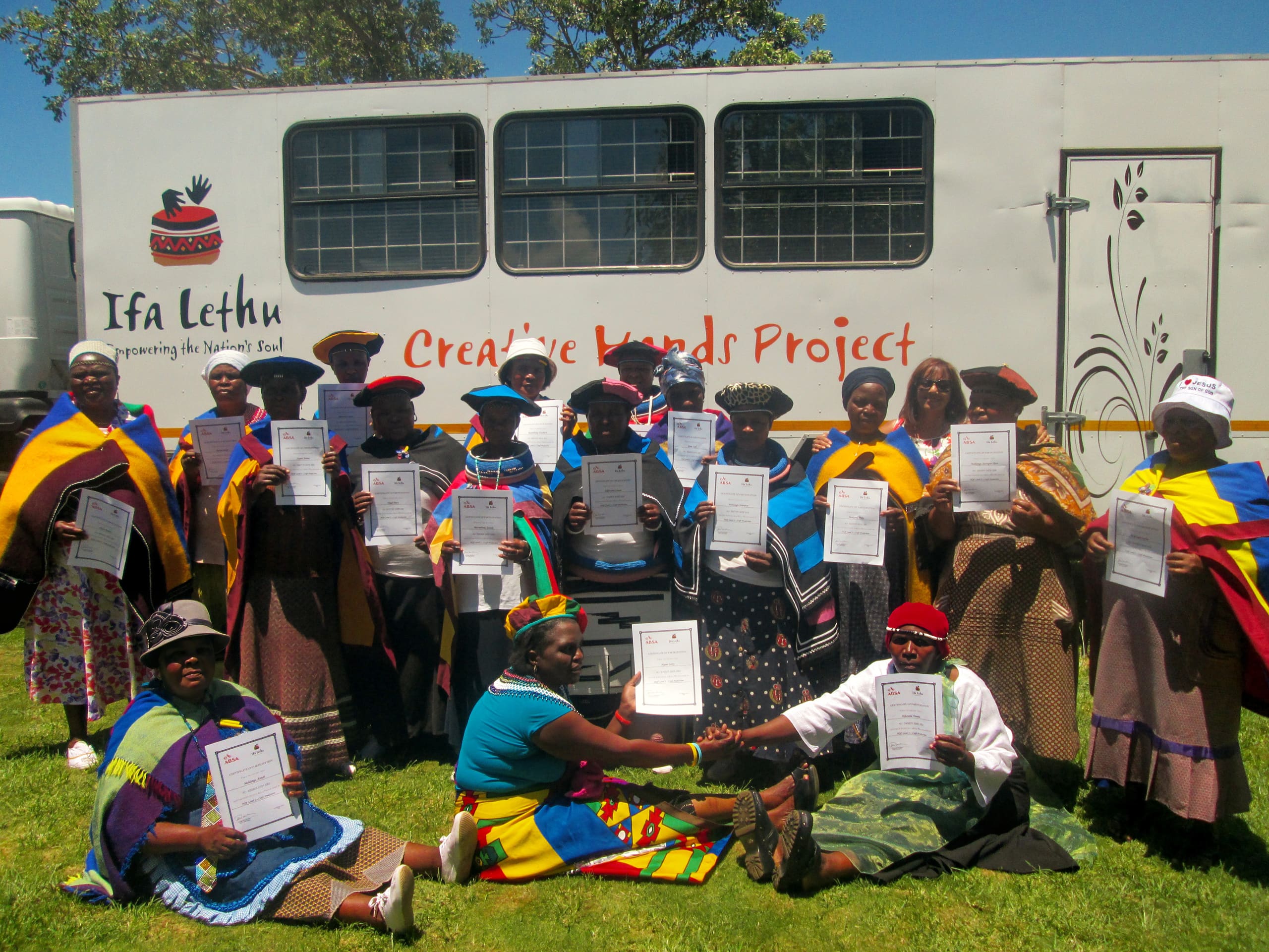 A group of people in colourful traditional attire hold certificates and pose in front of a white trailer with “Ifa Lethu Creative Hands Project” written on it. Some are seated on grass, while others stand, all smiling under a sunny sky.
