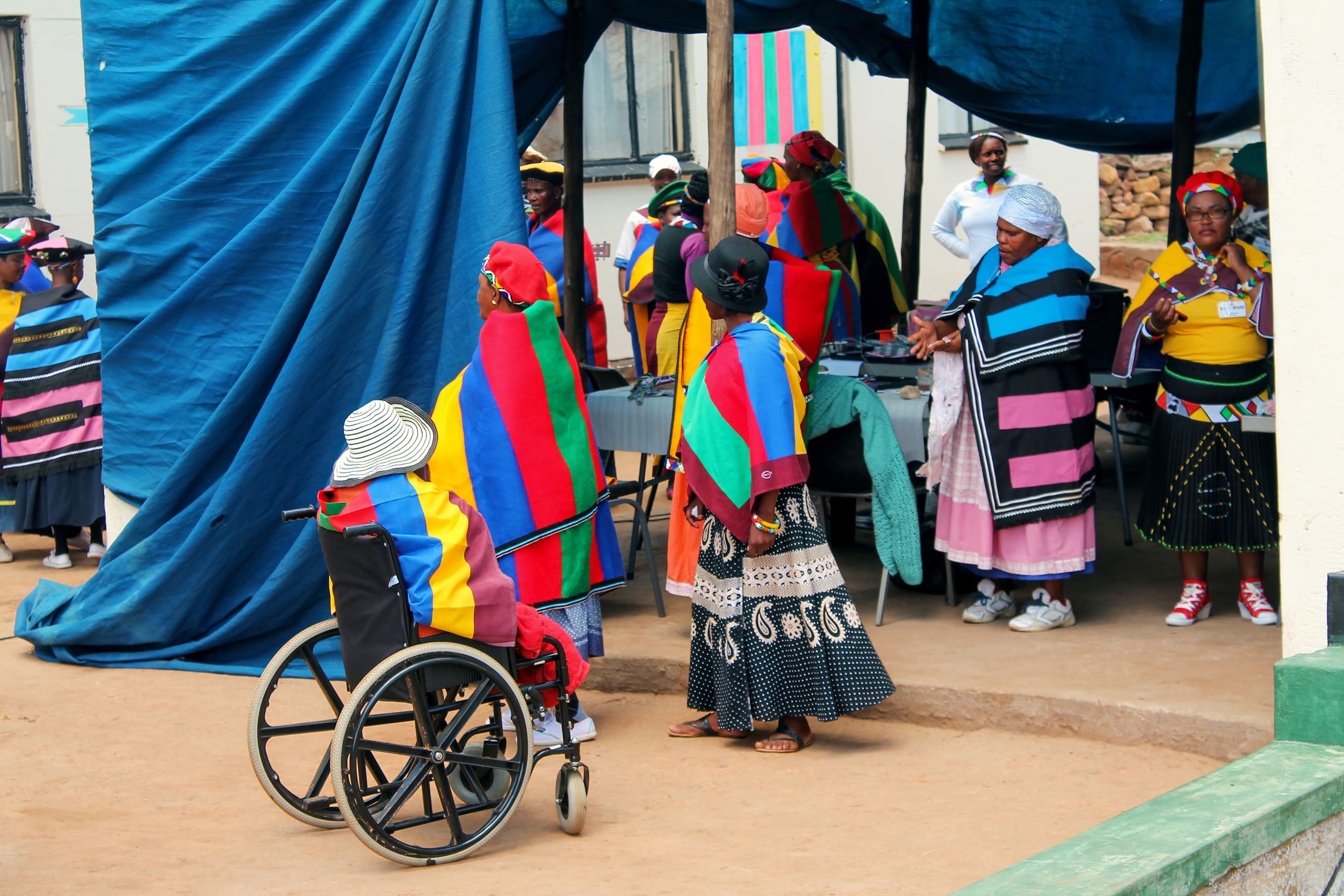 A group of women, some standing and one in a wheelchair, wear vibrant, striped traditional blankets and head wraps at an outdoor gathering under a blue canopy. The scene is colourful and lively, with women talking and engaging near tables set up outside.