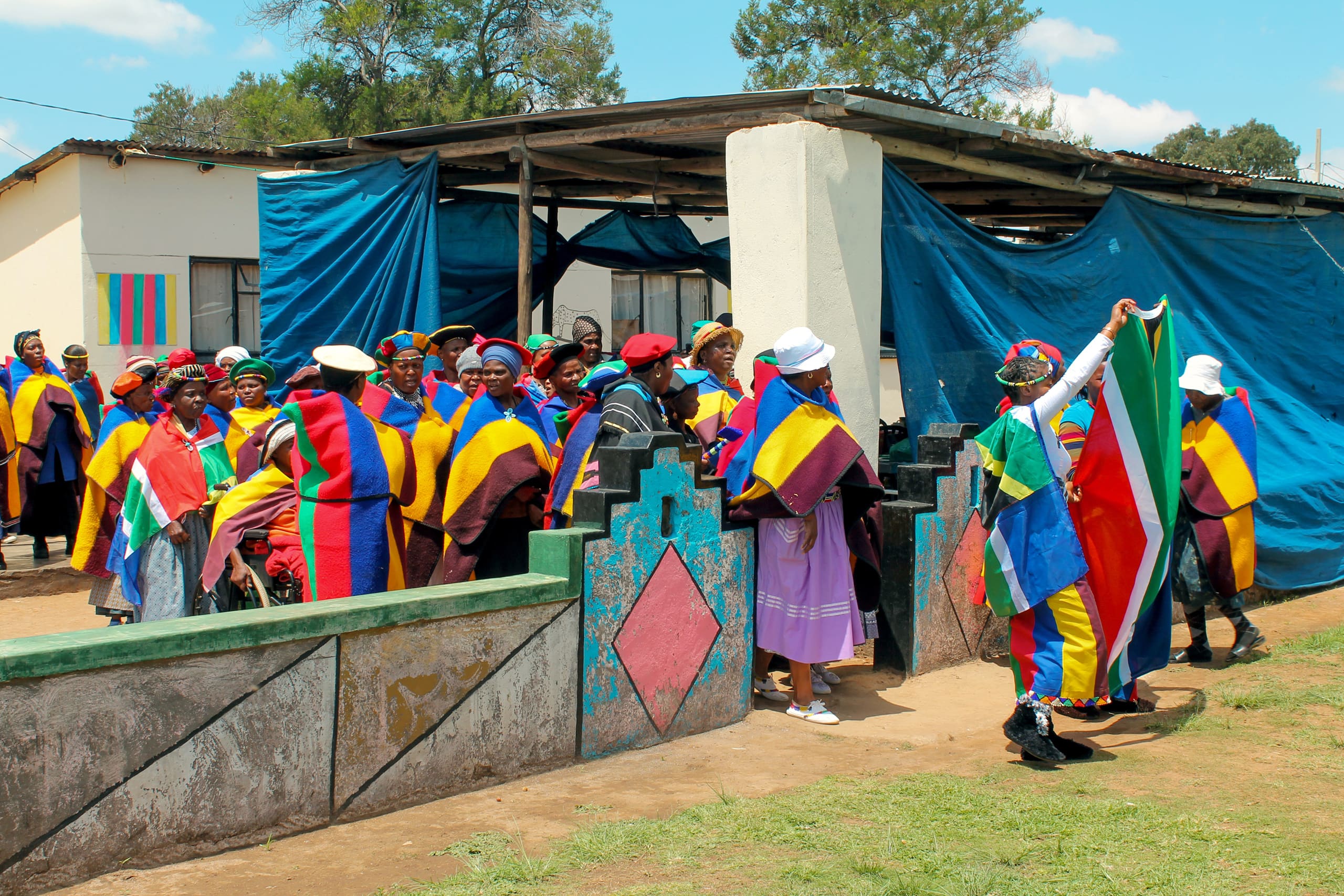 A group of people wearing colourful blankets and hats gather outside a house with blue tarpaulins, patterned walls, and painted decorations. One person holds a large South African flag, and the mood appears festive and communal under a clear sky.