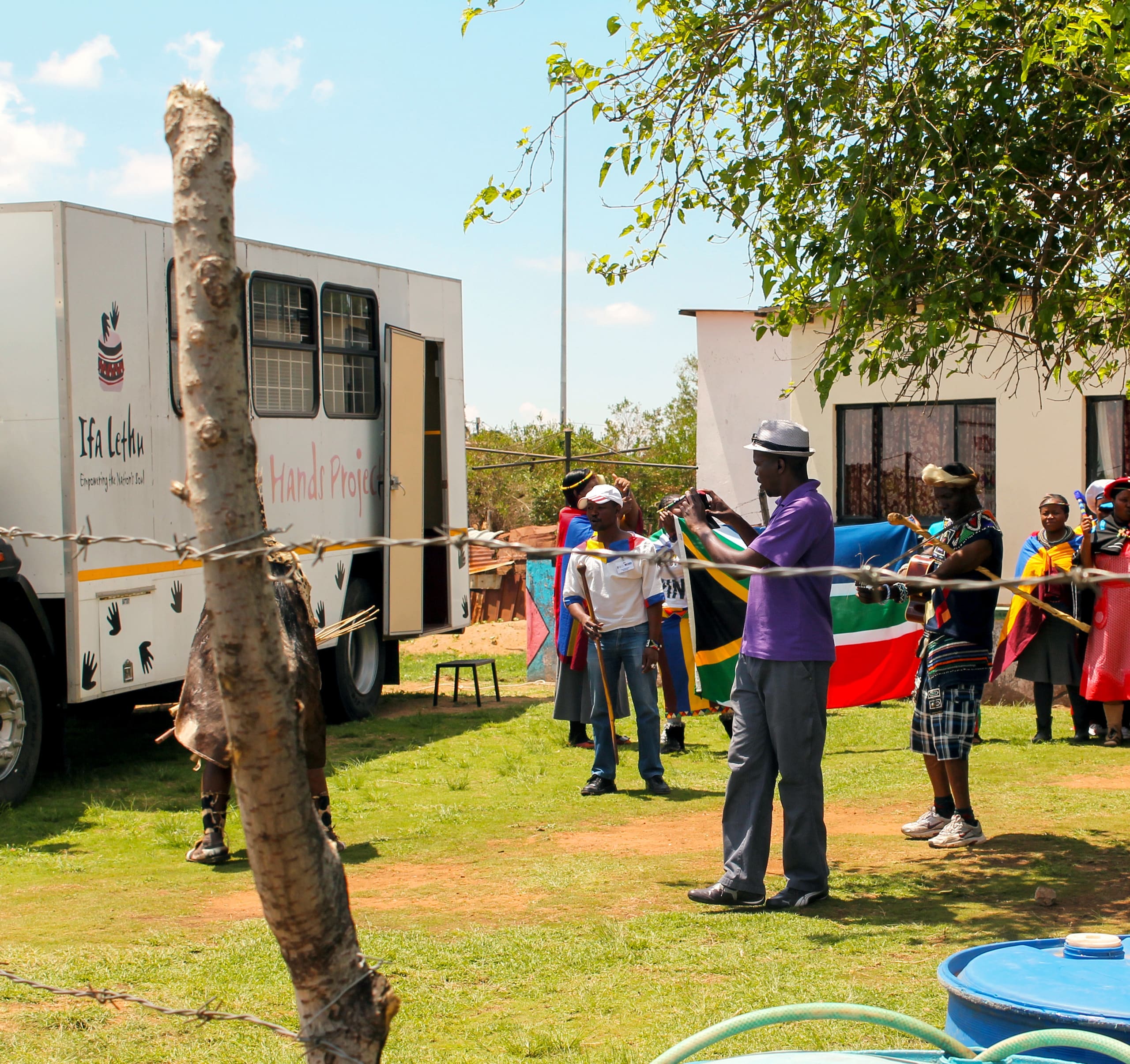 A group of people, some holding a South African flag, stand outside near a white lorry labelled 