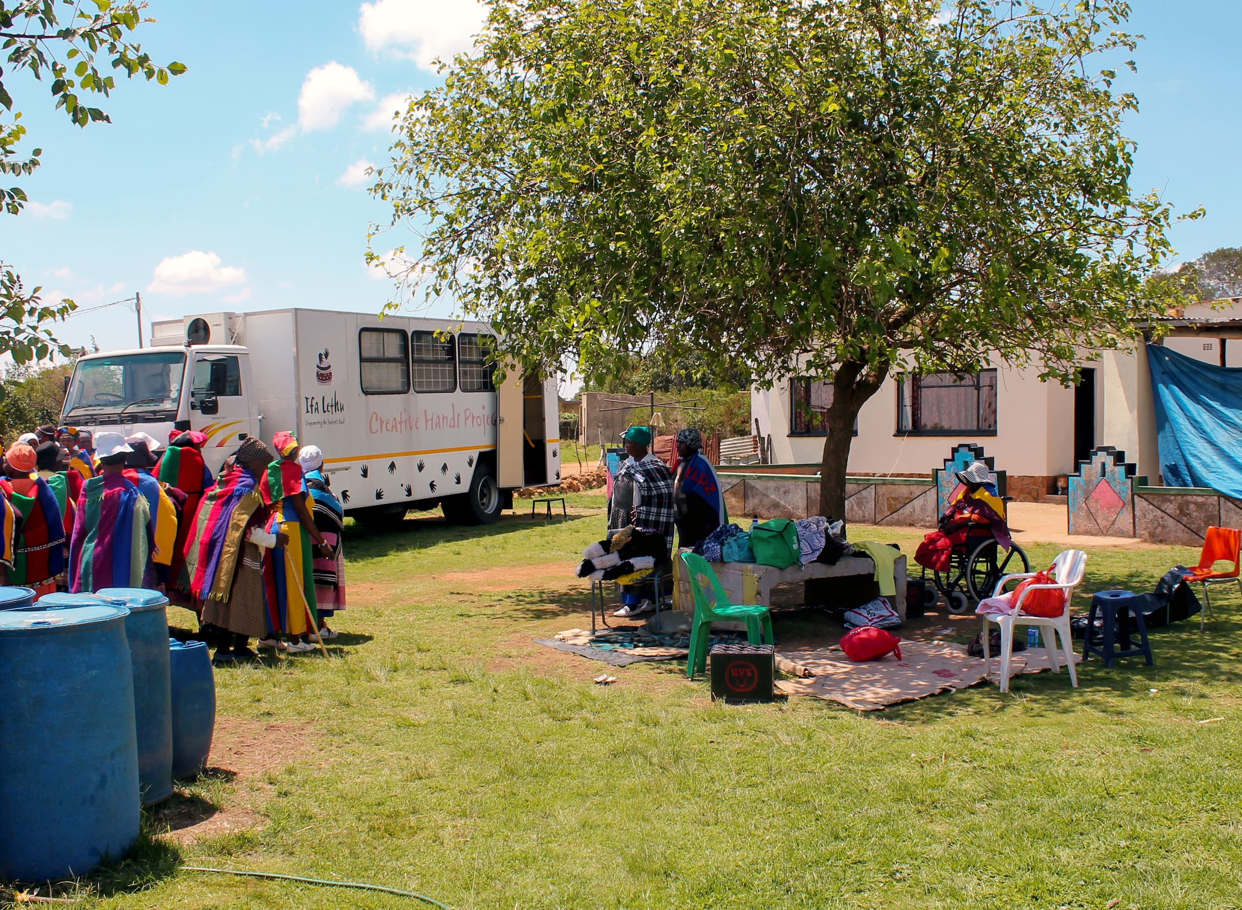 A group of women in colourful traditional clothing stands near blue barrels and a large white mobile clinic van parked on grass. Others sit under a tree, some on chairs, with supplies and bins nearby. A white building and a blue tarpaulin are in the background.