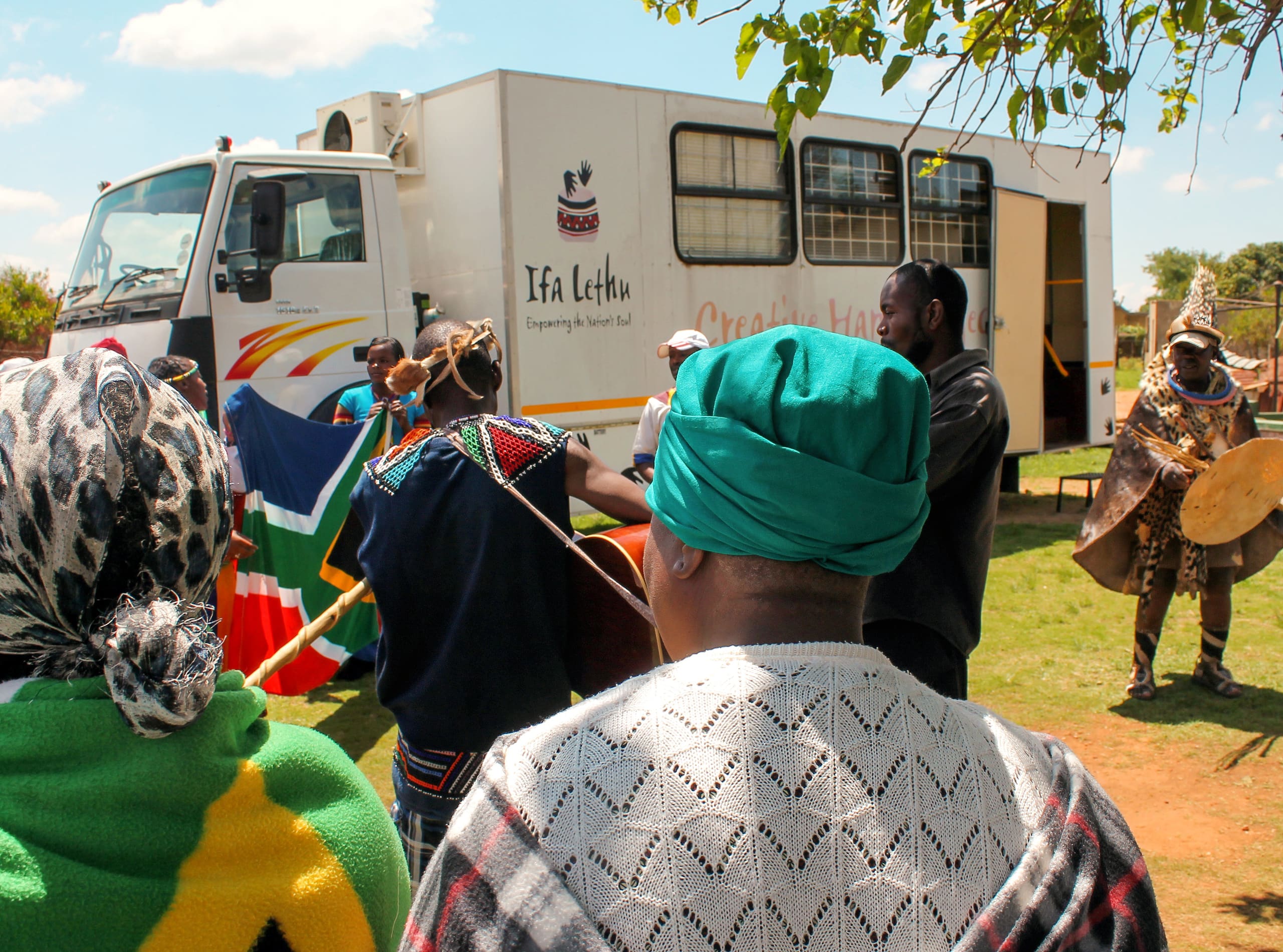 A group of people in colourful traditional African attire gather outdoors near a mobile museum lorry. Two women in headwraps stand in the foreground, while others hold a South African flag and musical instruments. The lorry is branded with 