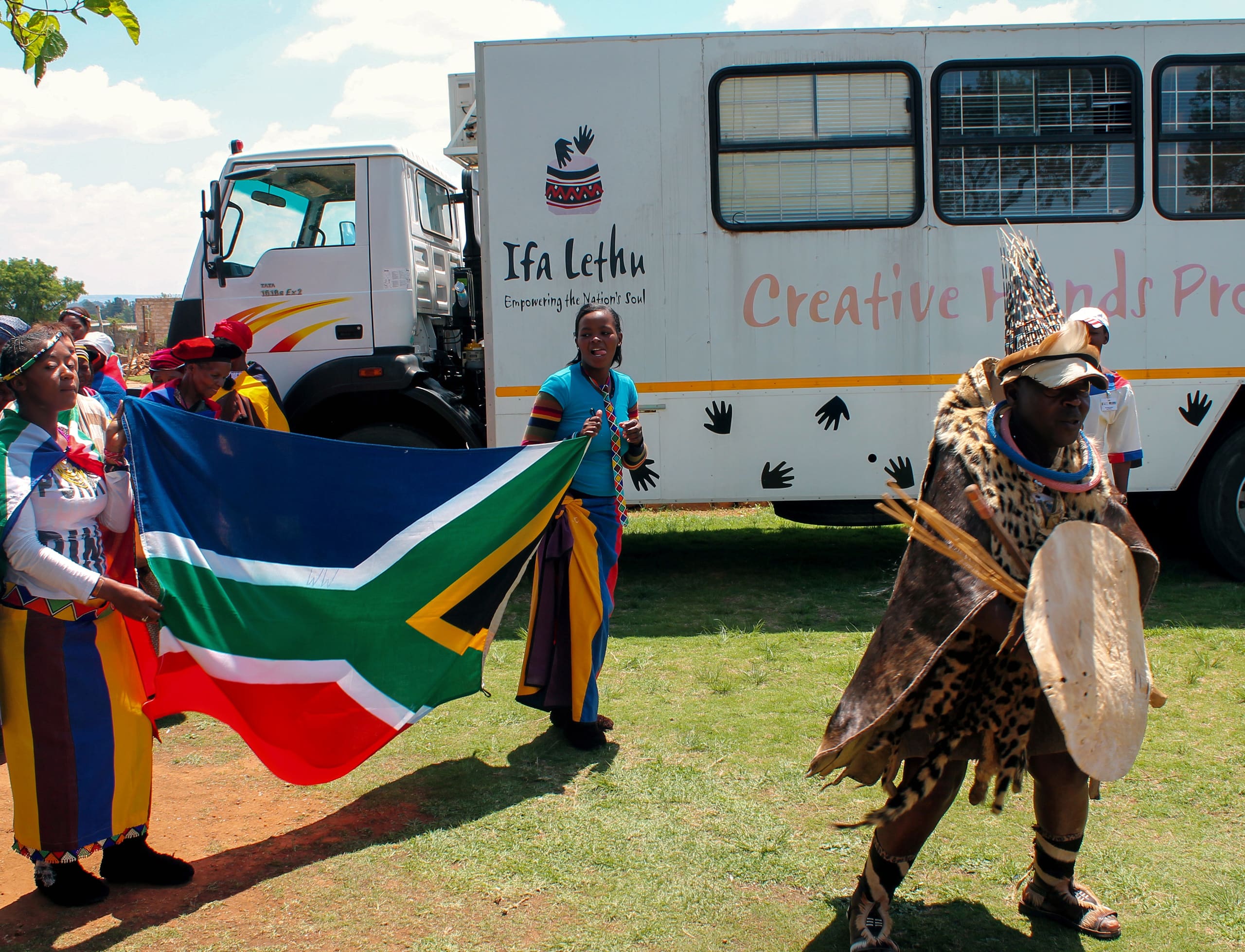 A group of people in colourful traditional attire stand outdoors, one holding a large South African flag. In front, a person dressed in animal skins and a headdress dances with a shield and staff. Behind them is a lorry with “Creative Hands Project” written on the side.