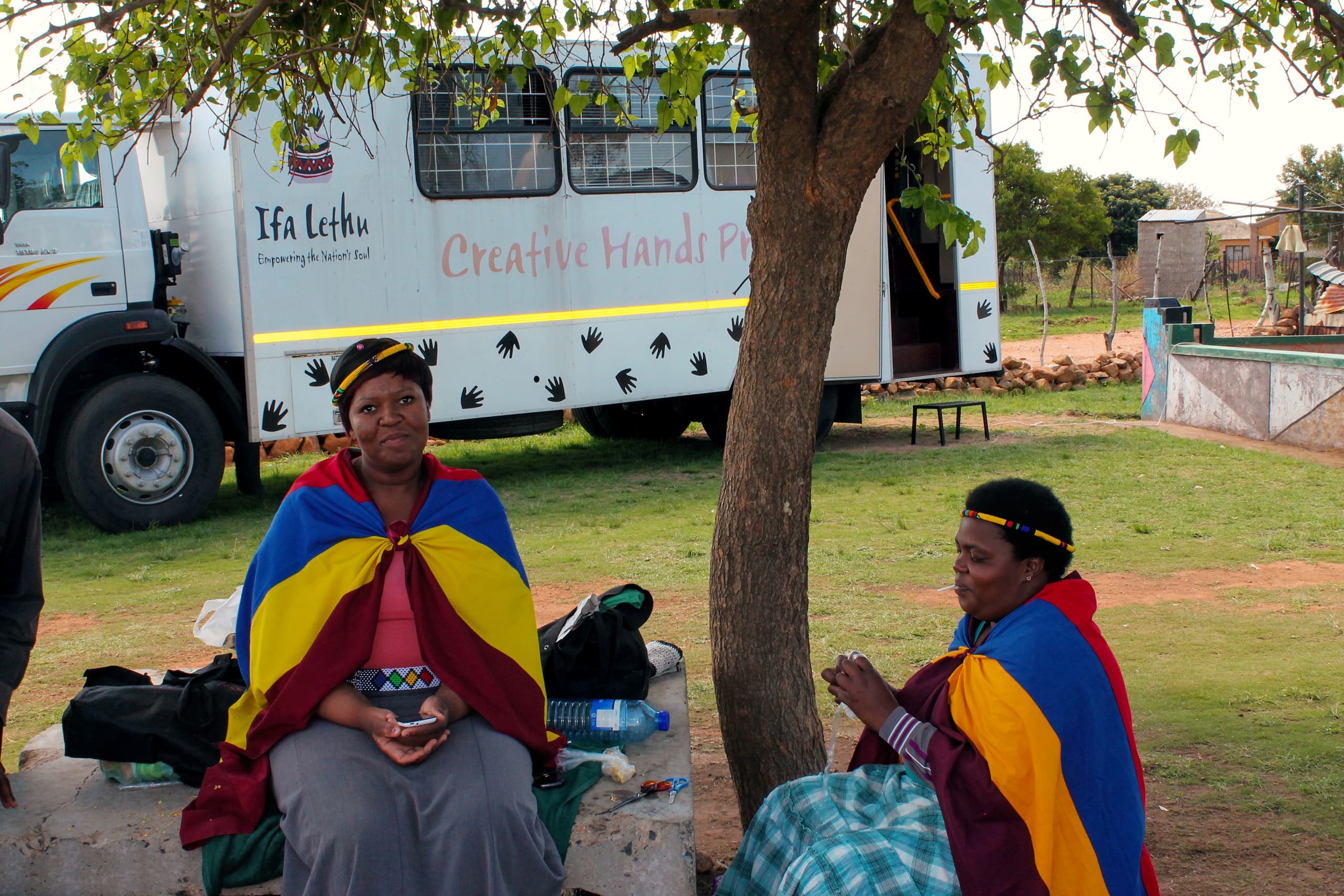 Two women sit under a tree, wearing brightly coloured traditional shawls in blue, yellow, and red. Behind them is a large white coach with 