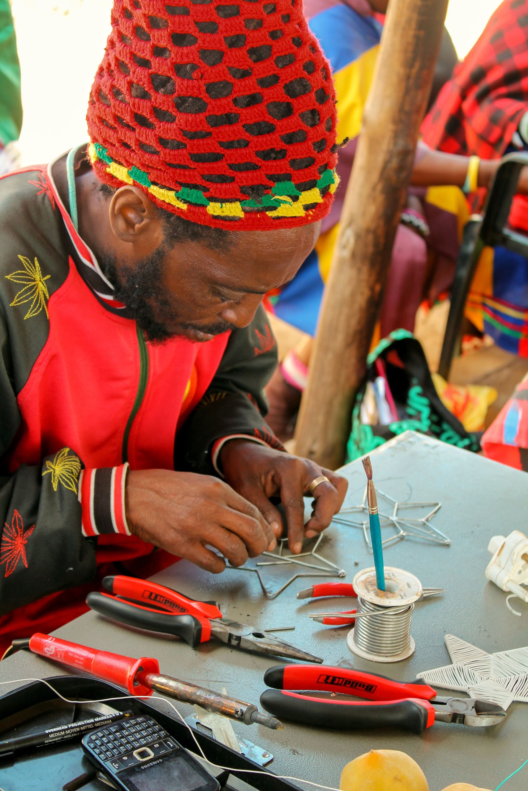 A man with a red, yellow, and green crocheted hat and a colourful jacket sits at a table, carefully crafting metal wire into star shapes. Tools, wire, a spool of solder, a brush, and a calculator are scattered on the table. Other people are visible in the background.