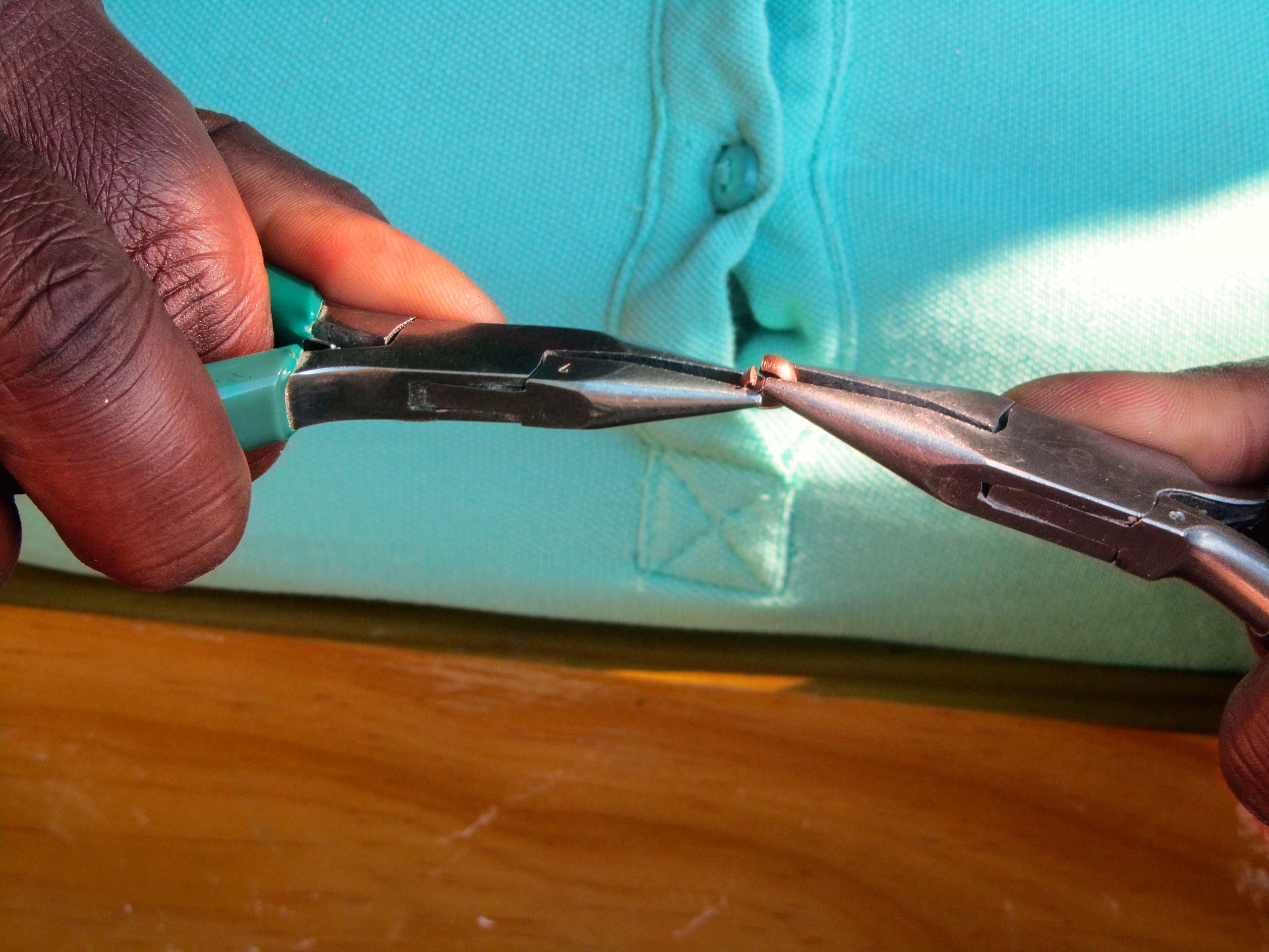 Close-up of two hands using pliers and long-nosed pliers to bend a small piece of wire. The person wears a turquoise shirt, visible in the background. The scene is well-lit, with a wooden surface below.