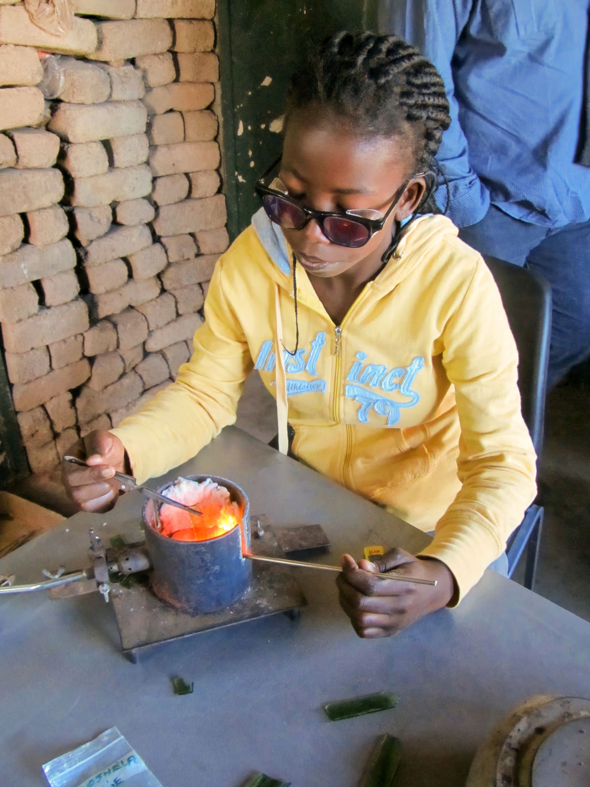 A person wearing dark safety glasses and a yellow hoodie carefully handles tools over a small metal furnace with a glowing hot interior, likely used for melting metal. Stacks of bricks are visible in the background and a packet lies on the table.