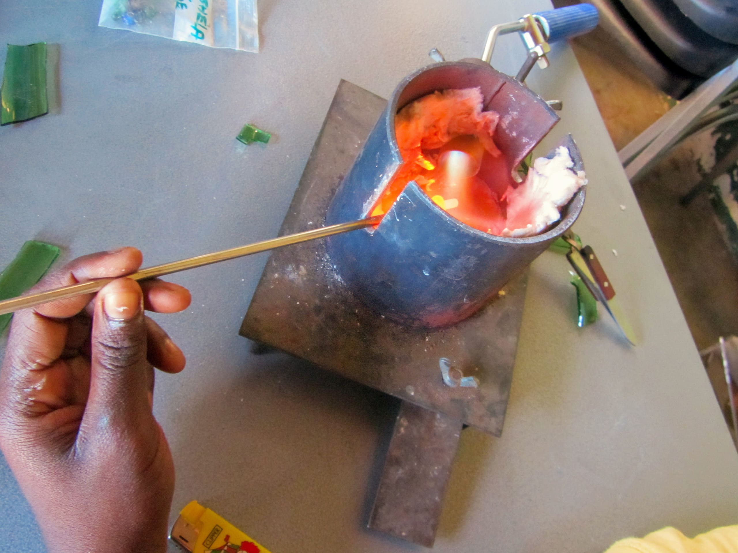 A hand holds a metal rod, stirring molten material in a cylindrical metal crucible with an open side. The crucible is heated and glowing orange inside, resting on a metal base. Green glass pieces, tools, and a yellow lighter are on the grey work surface.