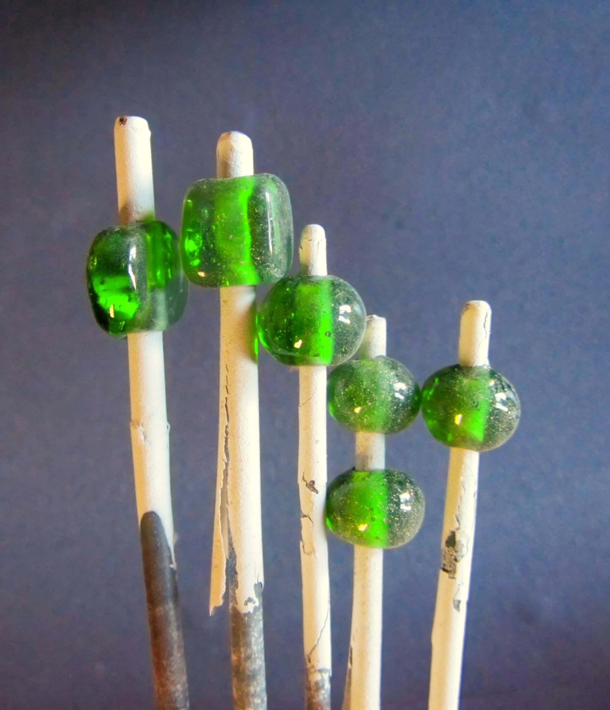 Five white rods with green glass beads on top stand against a plain blue background. The glass beads are transparent and slightly speckled, with two rods holding single beads and three holding two beads each. The rods are unevenly placed and a bit charred.