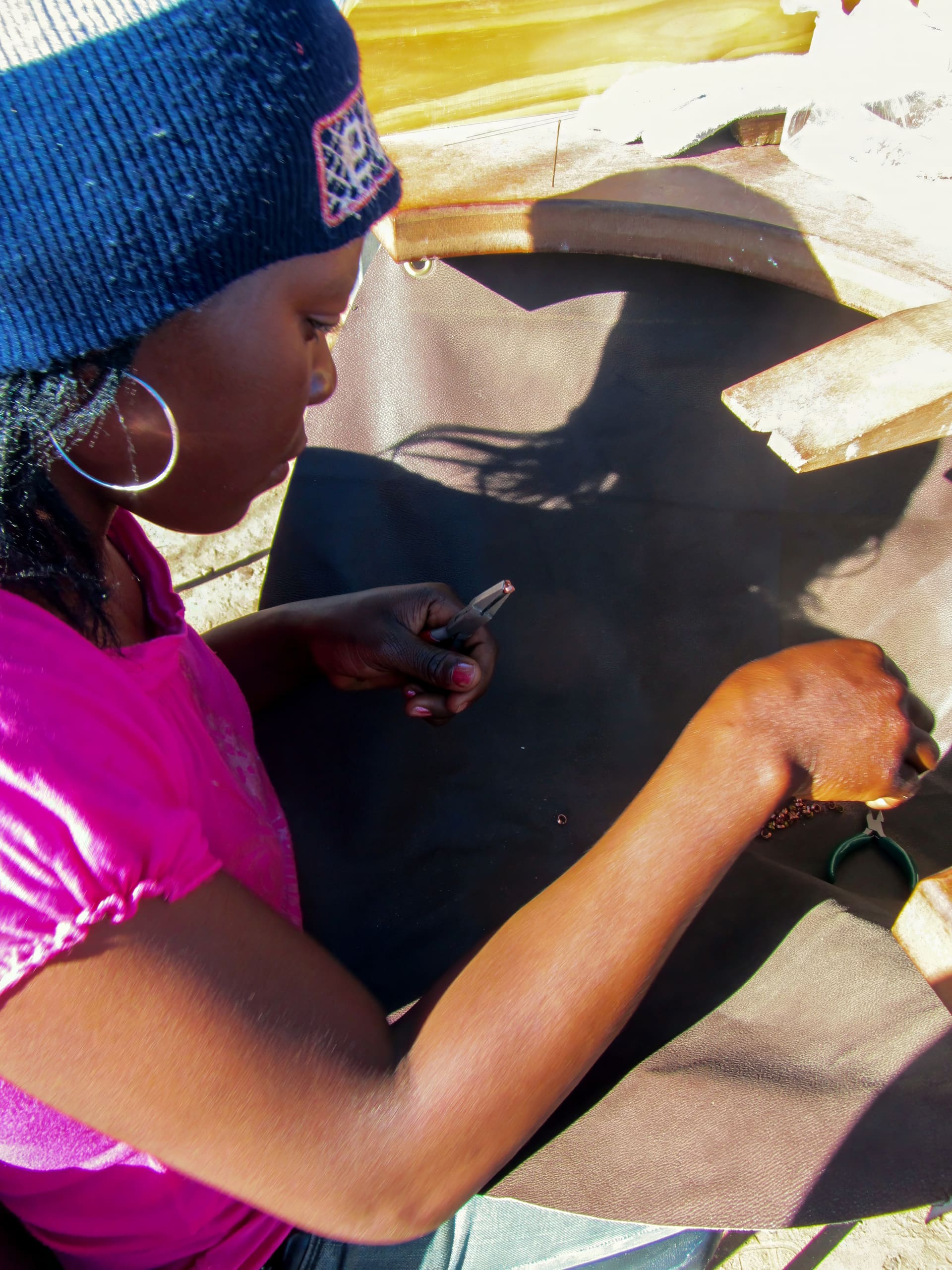 A woman wearing a blue knitted hat and pink shirt sits at a table, focused on creating jewellery. She holds small tools and carefully works with beads or wire on a dark surface. Sunlight casts shadows of her hands and head onto the table.